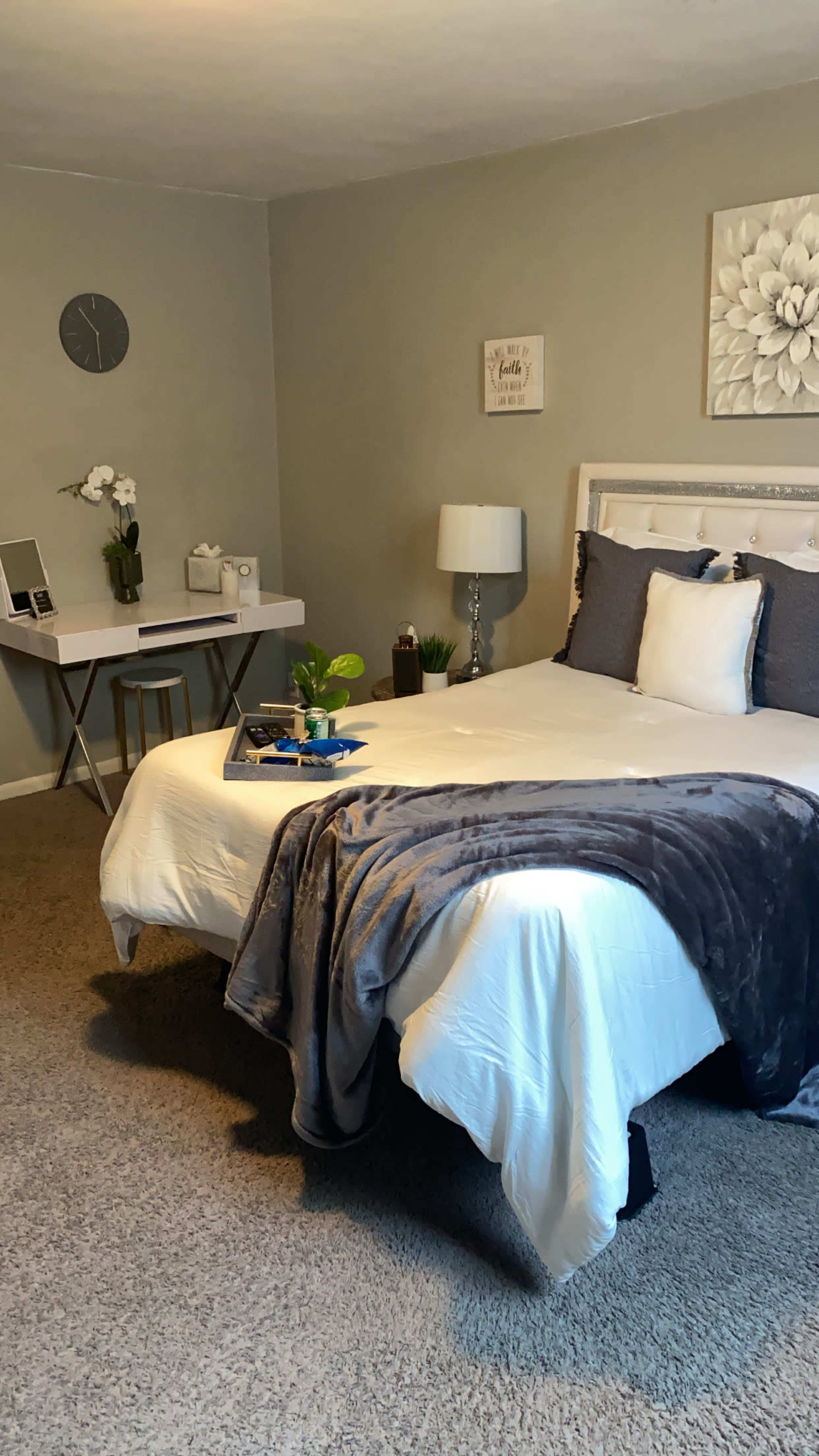 The image shows a neatly arranged bedroom featuring a bed with gray and white bedding, a small desk with a plant and decorative items, and a lamp beside the bed.