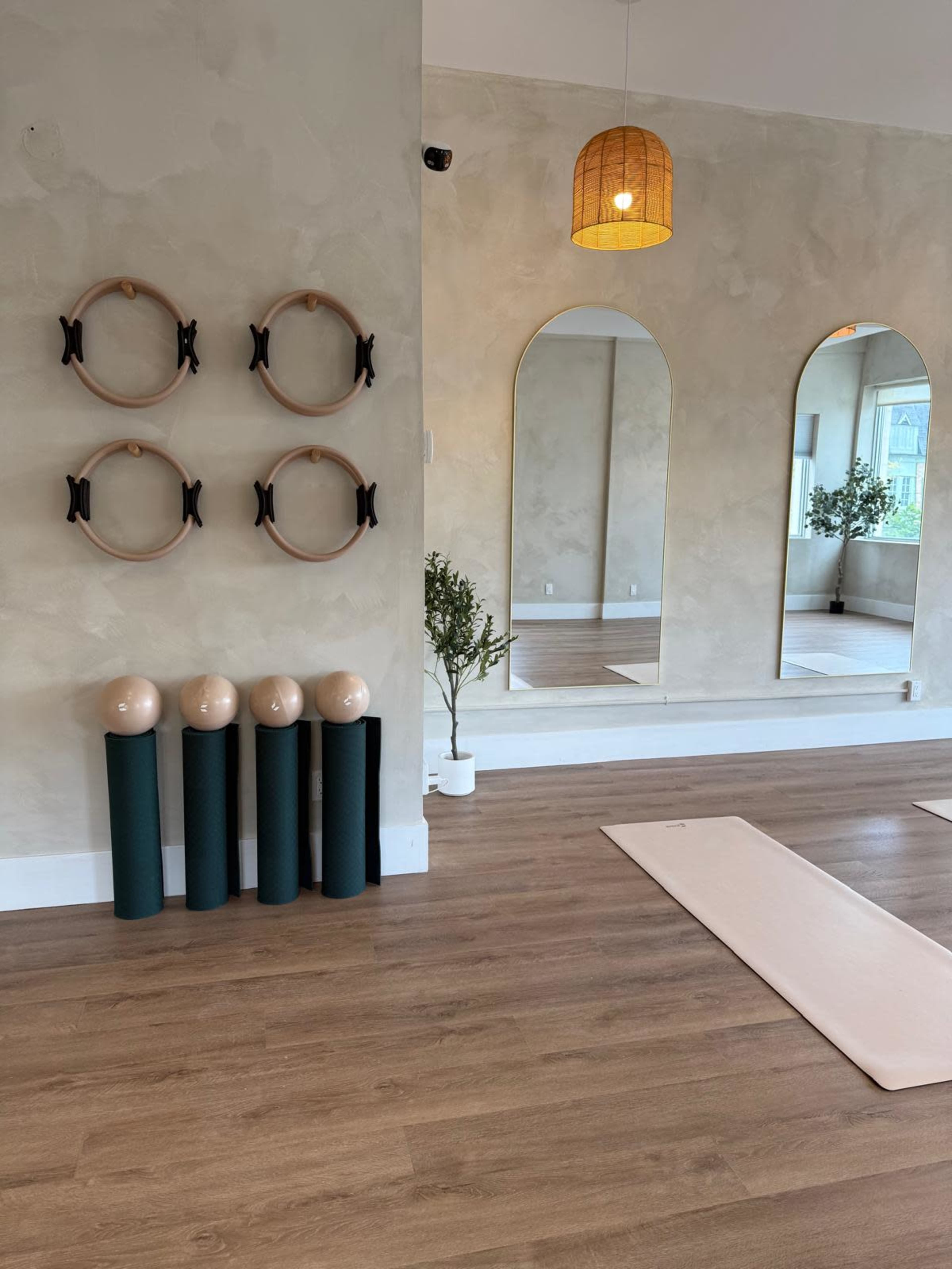 The image shows a minimalist exercise studio featuring four wall-mounted rings, three yoga mats rolled up in vertical holders, and two mirrors reflecting the light from a pendant lamp.