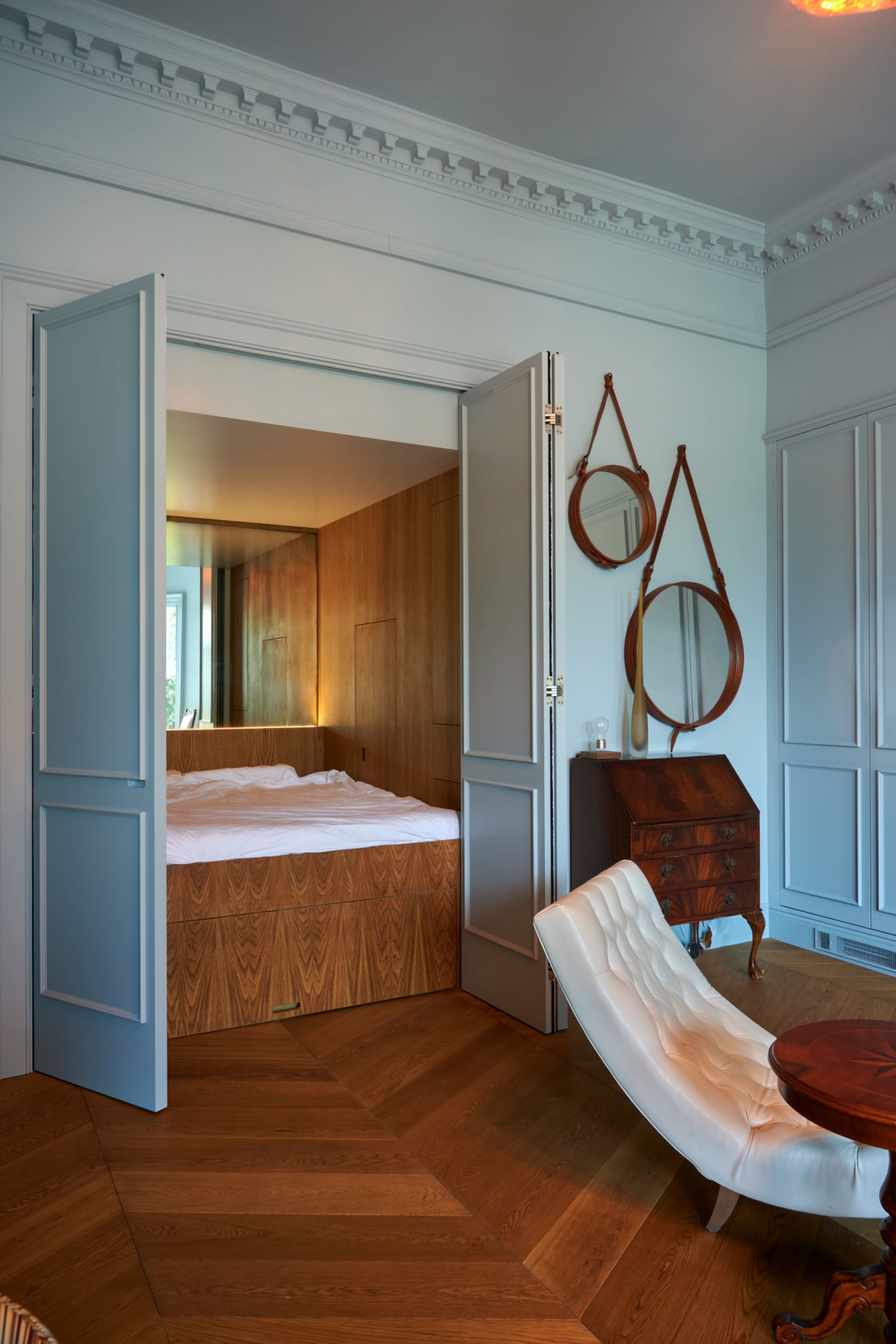 A bedroom is visible through open double doors, featuring a wooden platform bed and contrasting decorative elements in a softly lit modern interior.