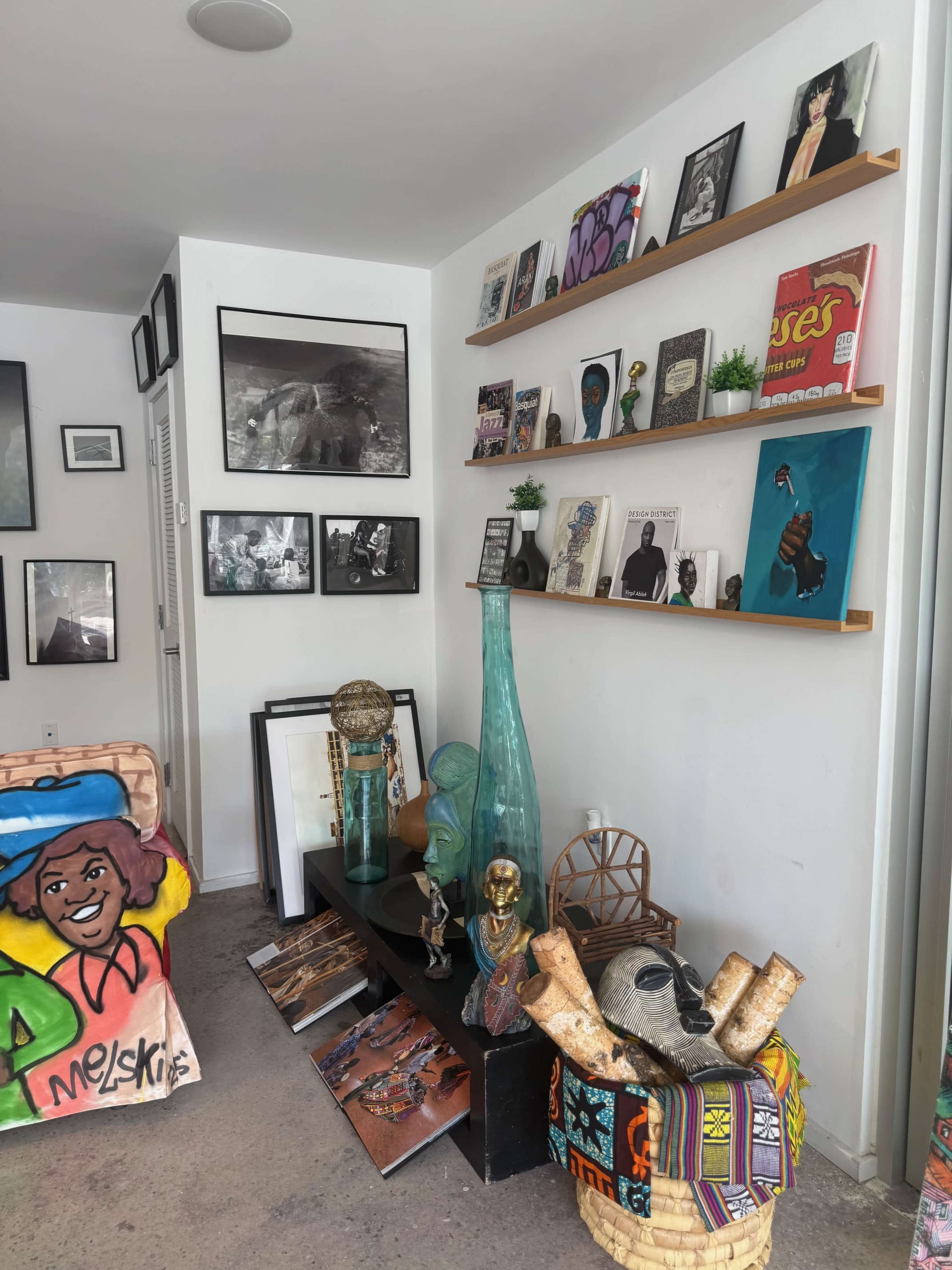 The image shows a corner of a room featuring a shelf filled with various framed artworks and decorative items, alongside a dark table displaying sculptures and colorful art pieces.