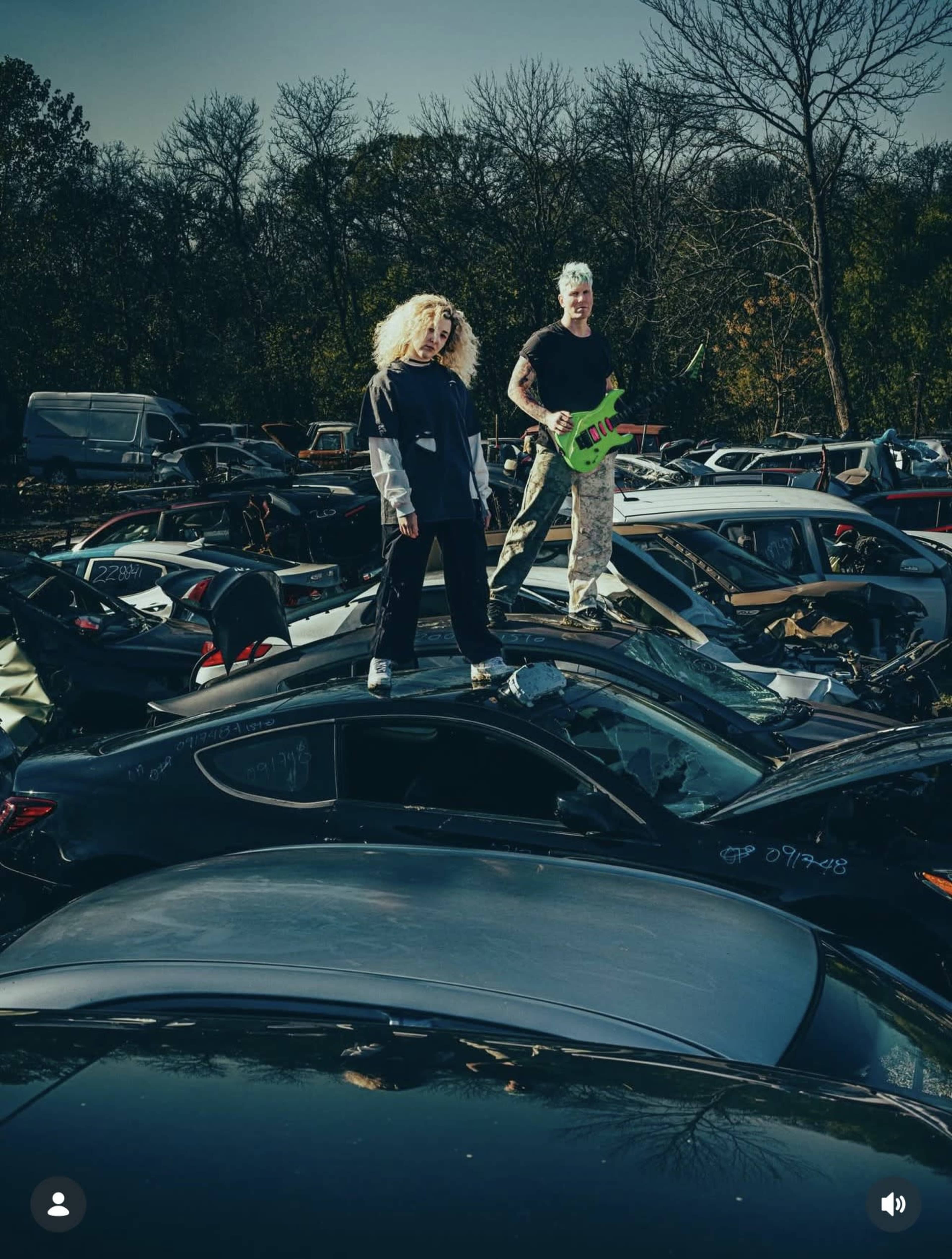 Two individuals stand on top of crushed cars in a junkyard, surrounded by various discarded vehicles under a clear sky.