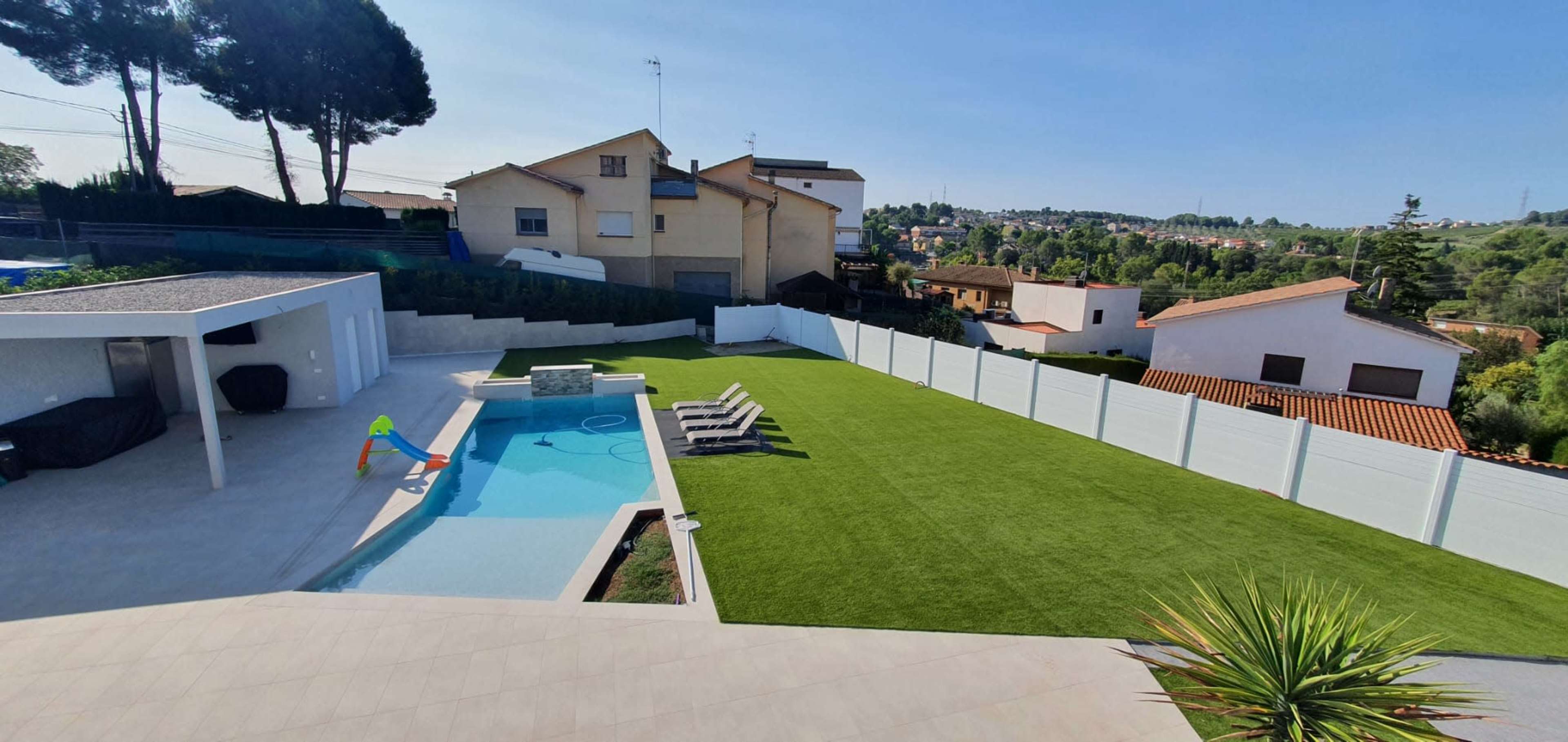 A backyard with a swimming pool, lounge chairs, and a well-maintained lawn, surrounded by white fences and residential buildings in the background.
