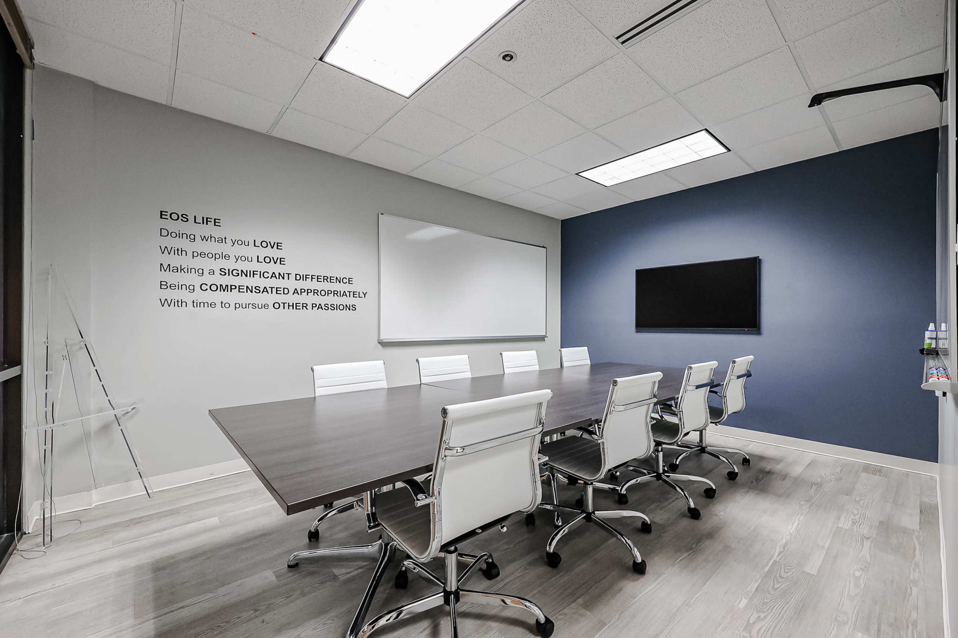 A modern conference room features a long wooden table surrounded by white chairs, a large wall-mounted screen, and a whiteboard, with motivational phrases on the adjacent wall.