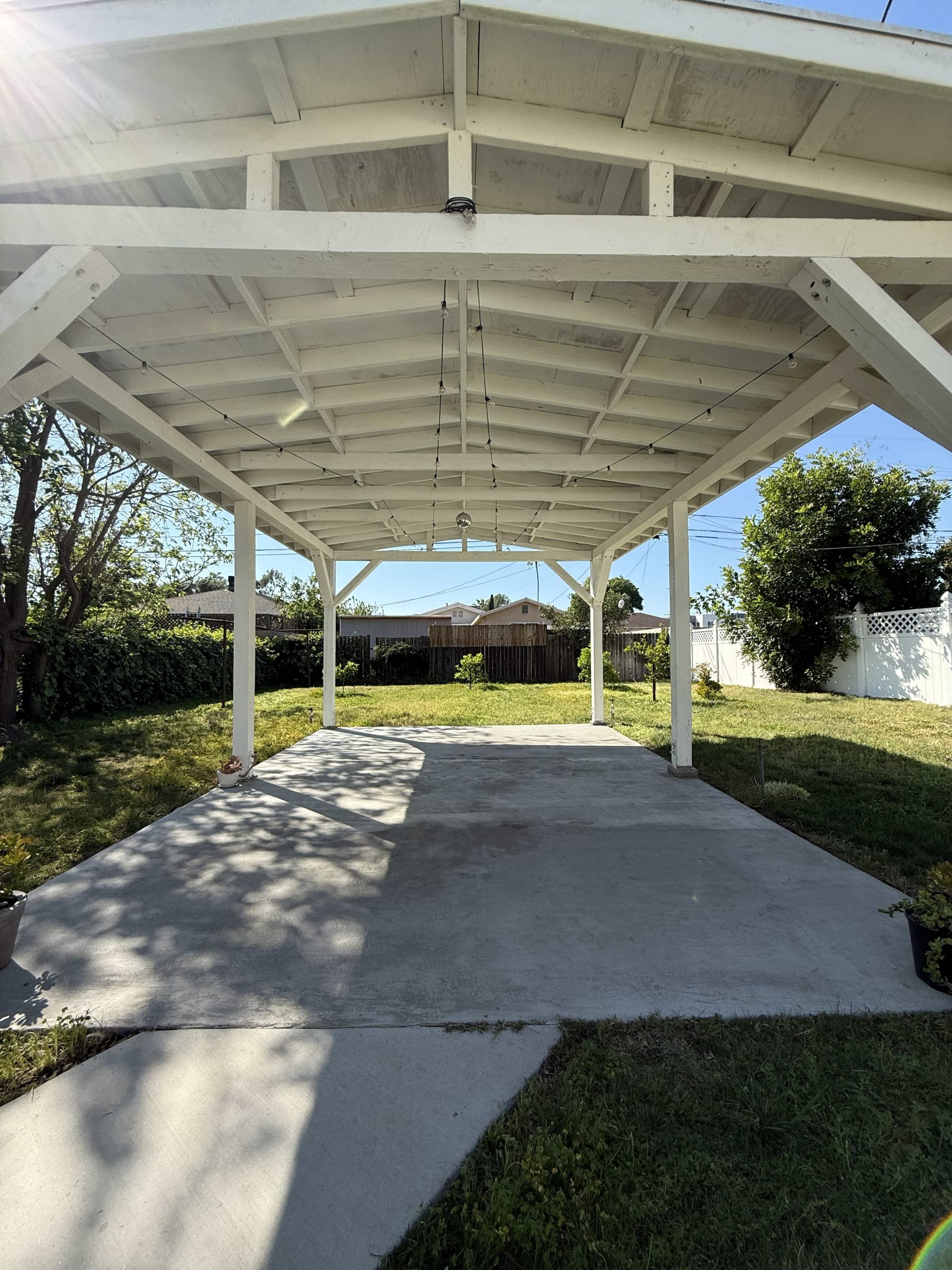 The image shows a spacious, covered patio with a concrete floor, surrounded by grass and trees in a backyard setting.