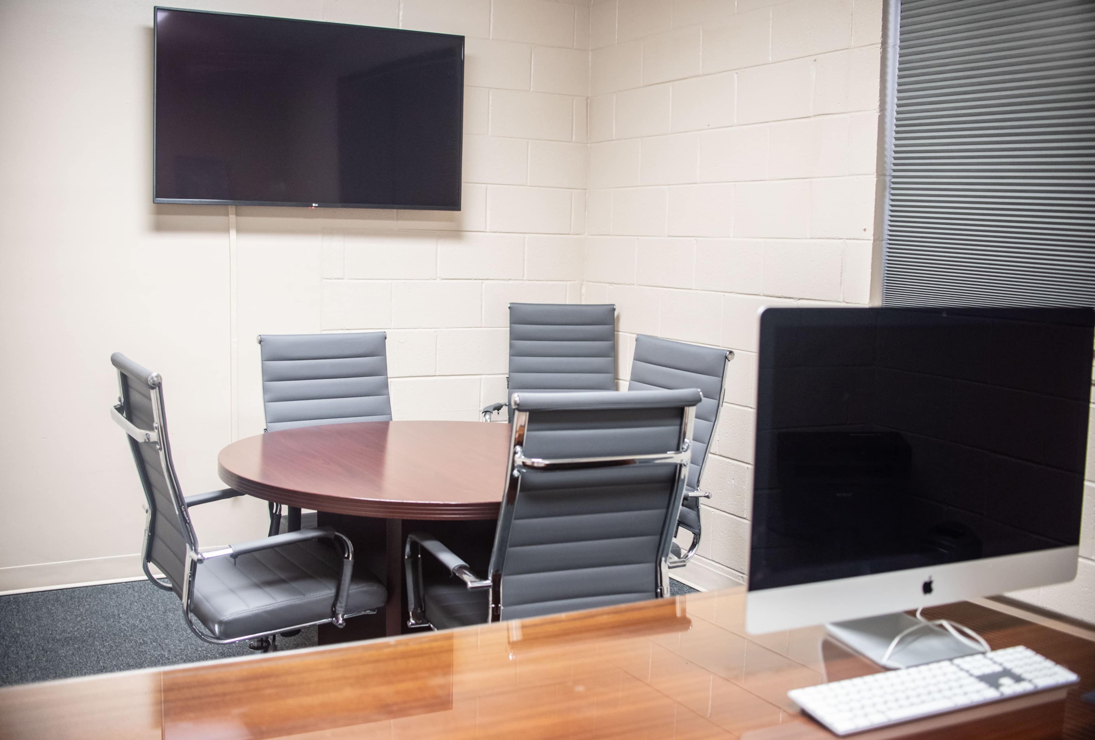 A conference room features a round table surrounded by five chairs, a wall-mounted television, and a computer on a desk.
