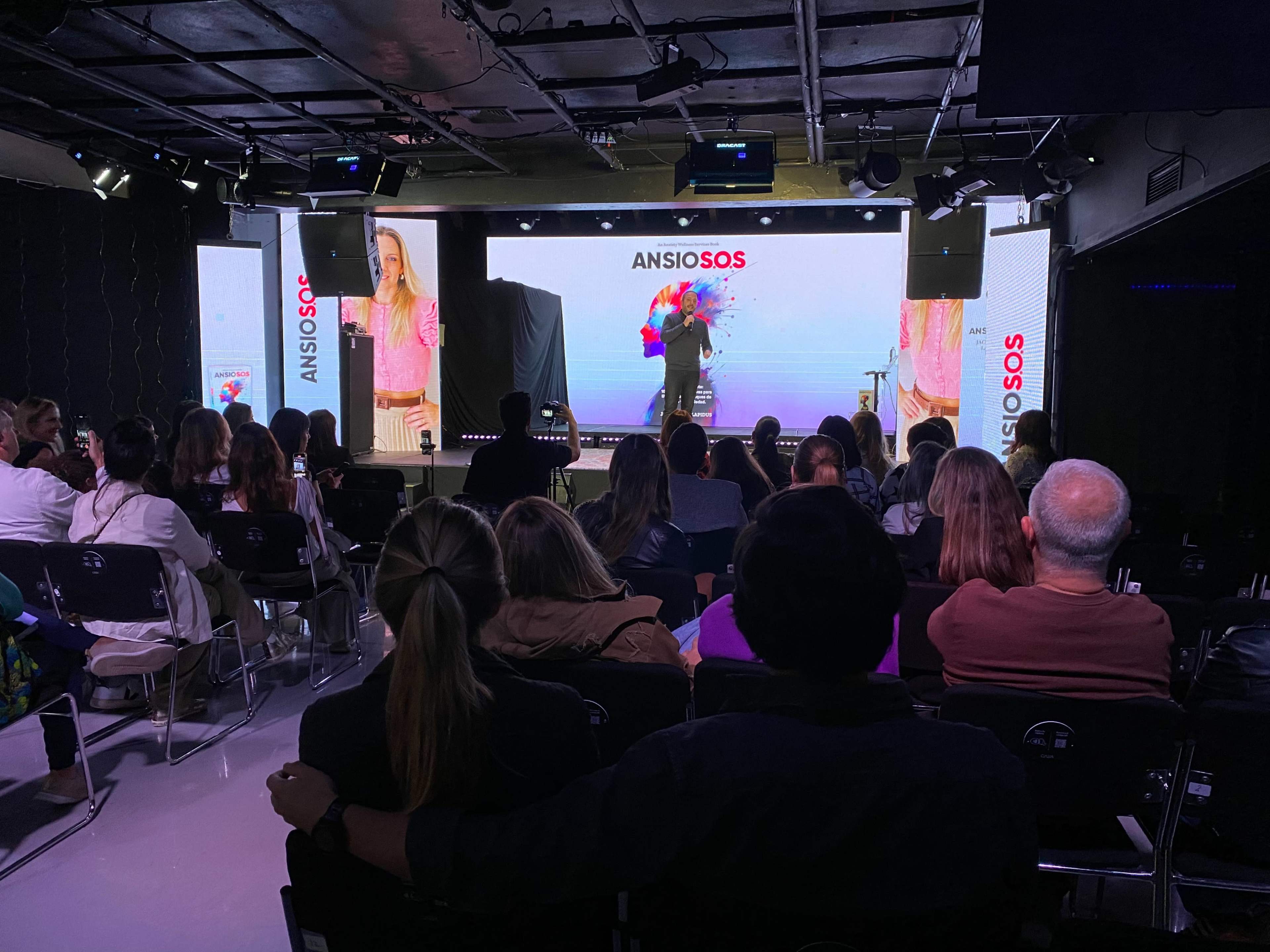A diverse audience sits in rows facing a large screen displaying a colorful presentation titled "ANSIO SOS."