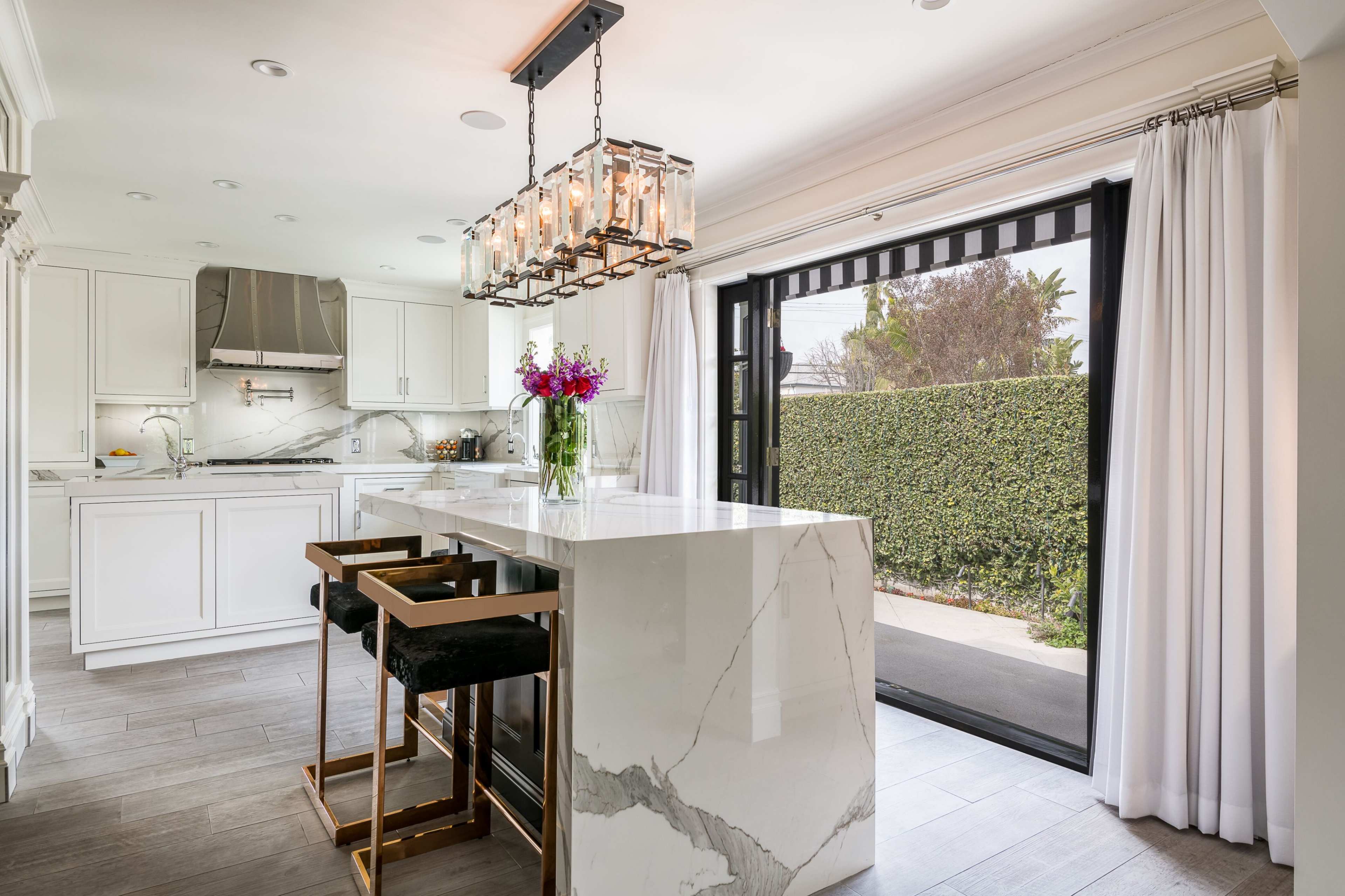 A modern kitchen features a large marble island with bar seating, bright natural light from a sliding glass door, and a chandelier overhead.