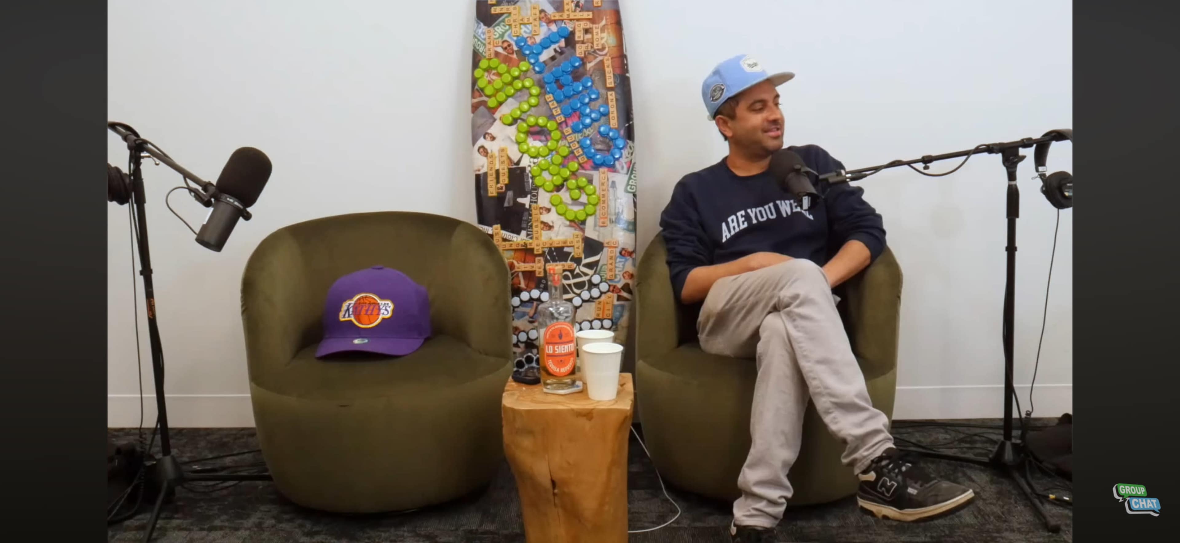 A man is sitting in a green chair, smiling, with a microphone in front of him, and a colorful skateboard mounted on the wall behind him.