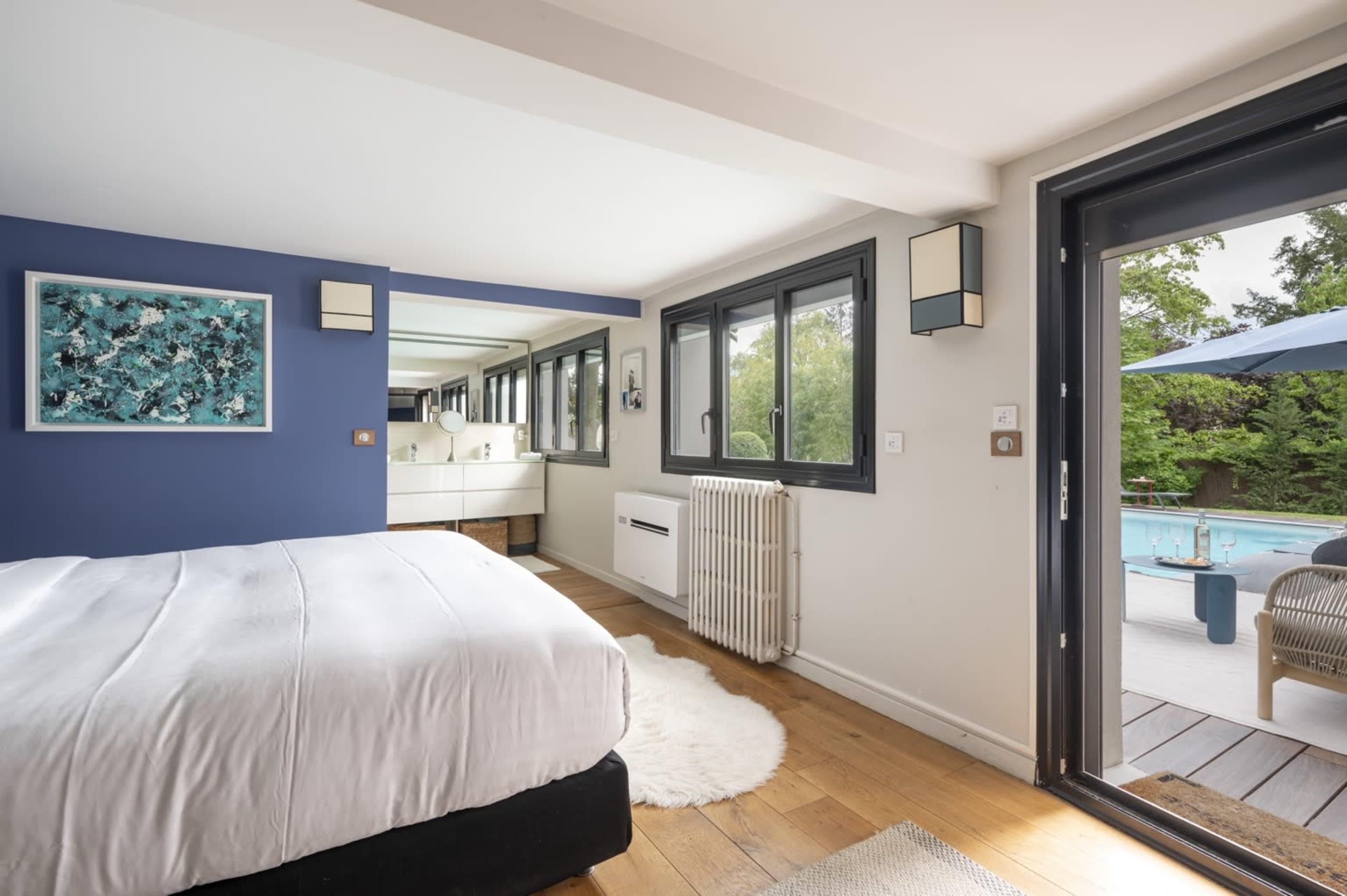 The image shows a modern bedroom with a large bed, wooden flooring, and a sliding glass door leading to an outdoor space with a pool.