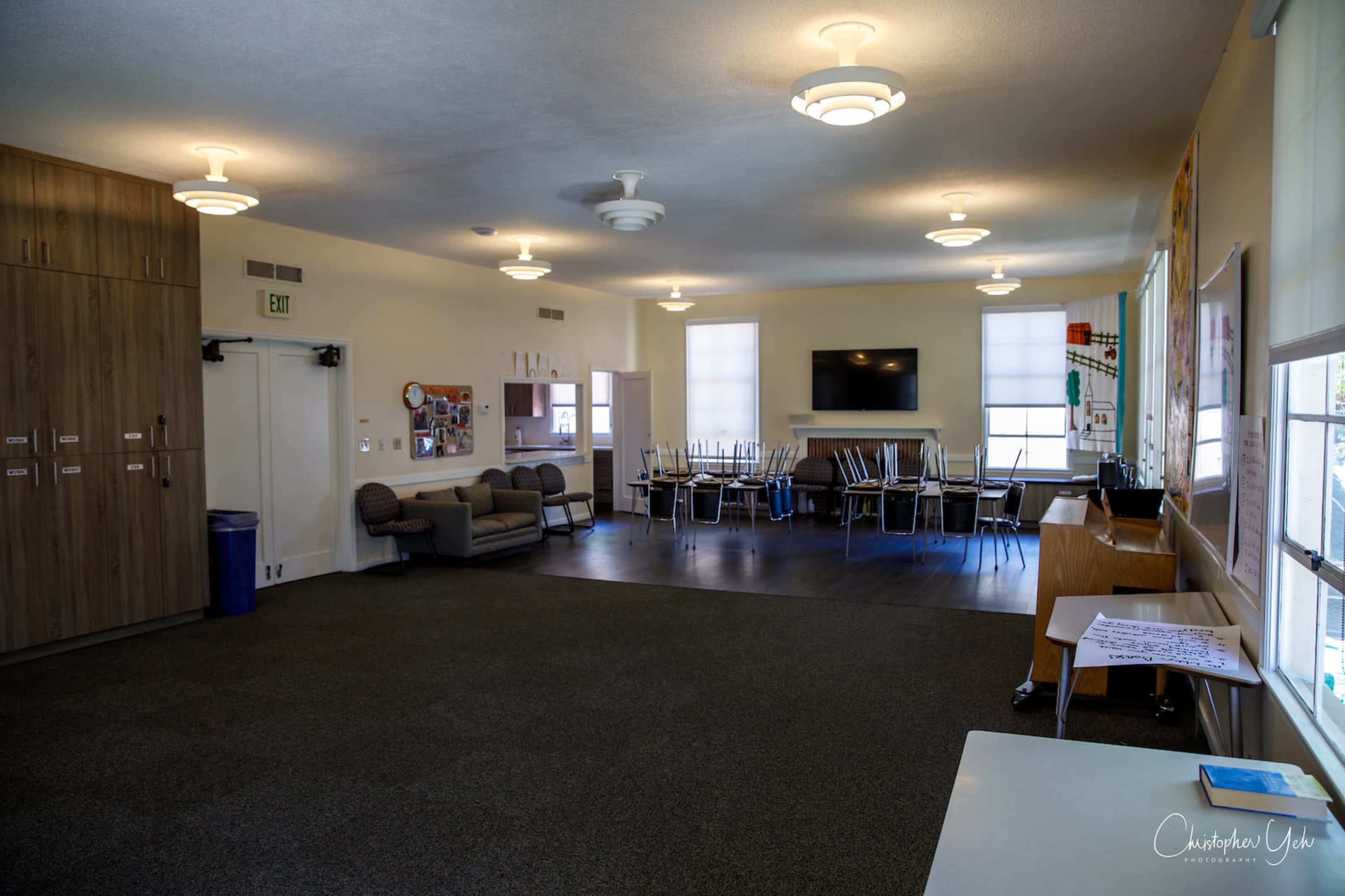 The image shows a spacious, well-lit room with a seating area, several tables and chairs, and a television mounted on the wall.