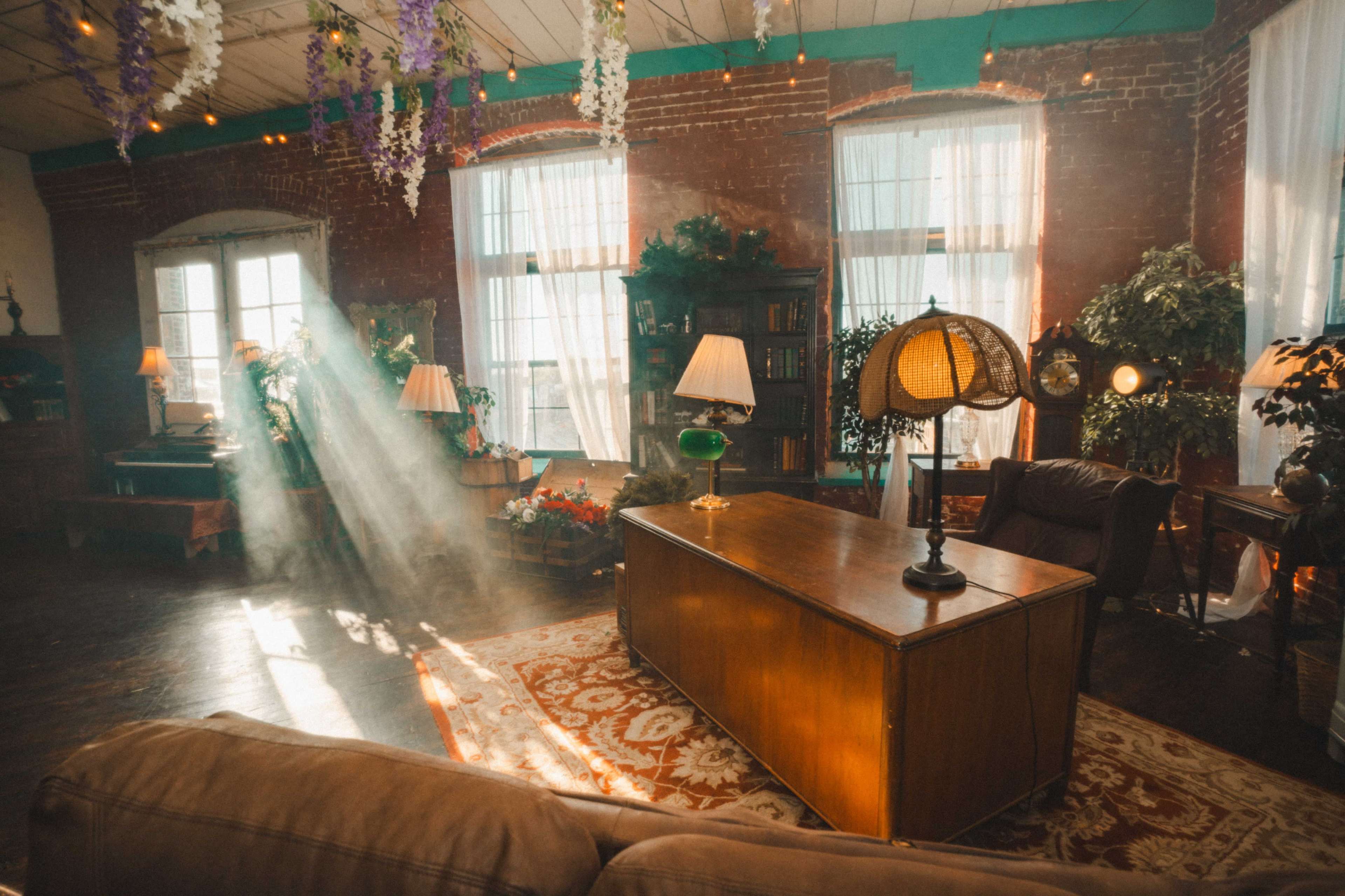 The image depicts a cozy room with large windows, a wooden desk, a sofa, and various houseplants, illuminated by soft rays of sunlight filtering through the curtains.