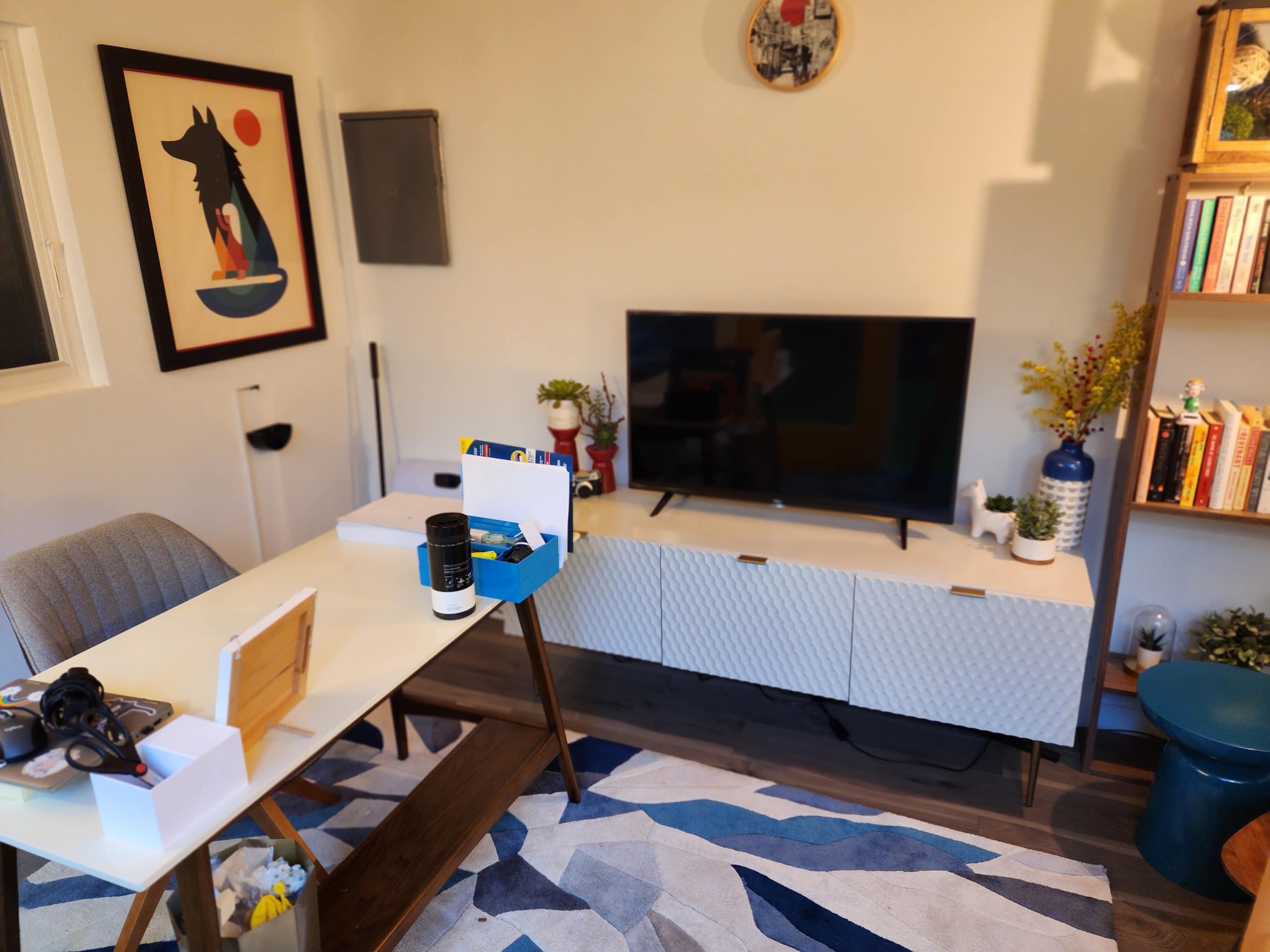 A modern home office is set up with a desk, a television on a low cabinet, a decorative artwork on the wall, and a rug on the floor.