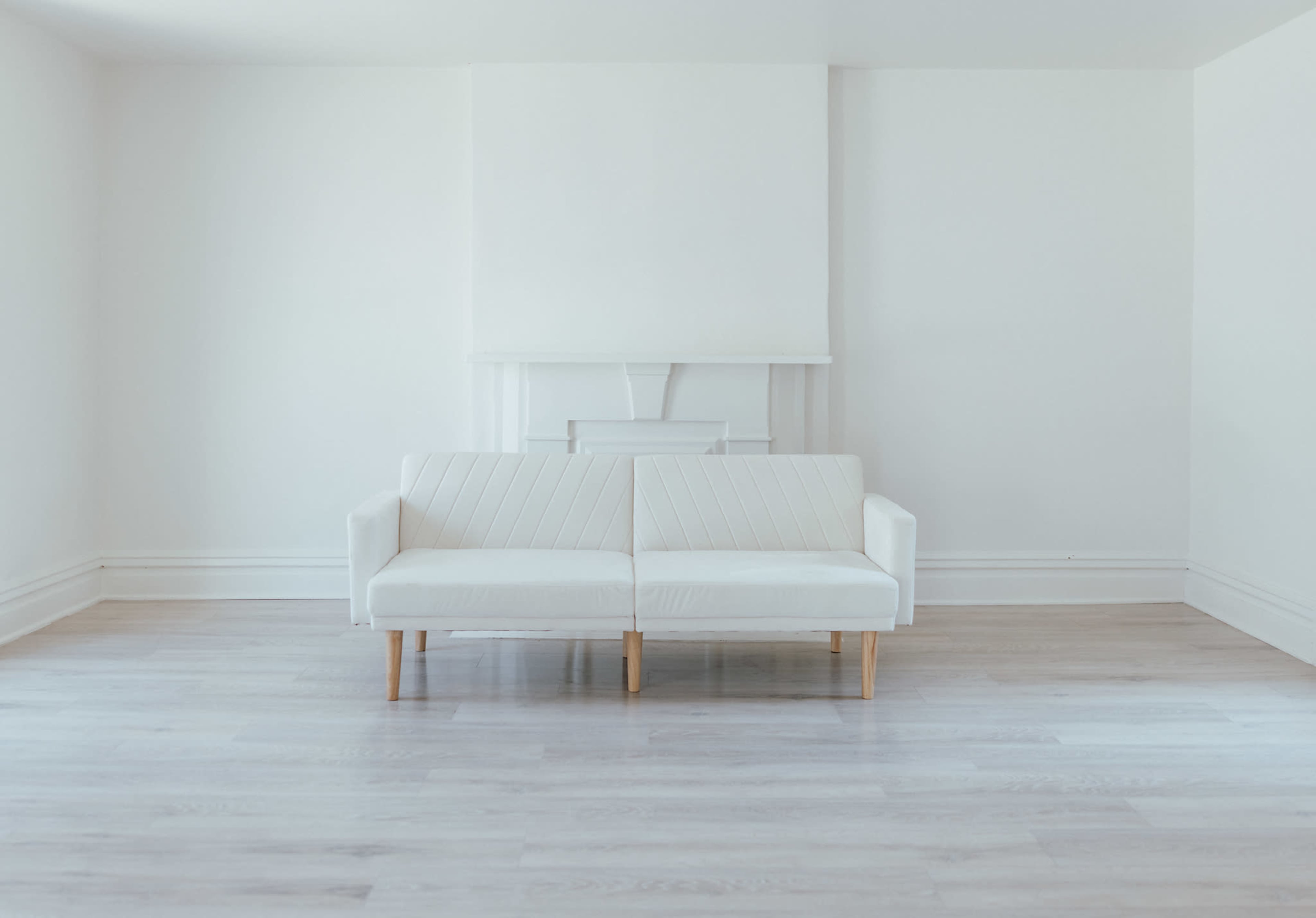 The image shows a white, modern couch positioned in a minimalistic room with light wooden flooring and white walls.
