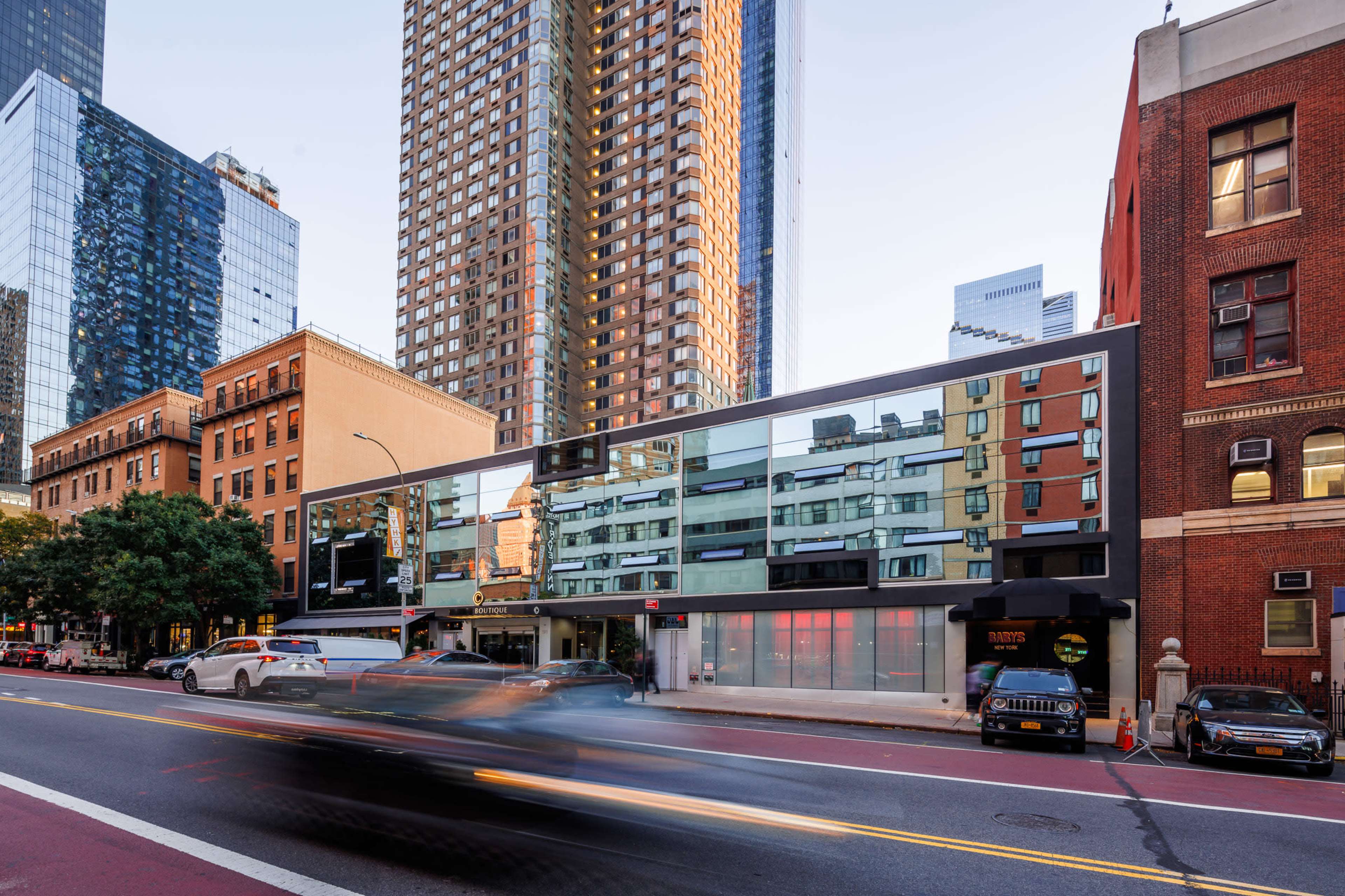 A modern building with large glass windows is situated on a city street lined with a mix of tall skyscrapers and older brick structures.