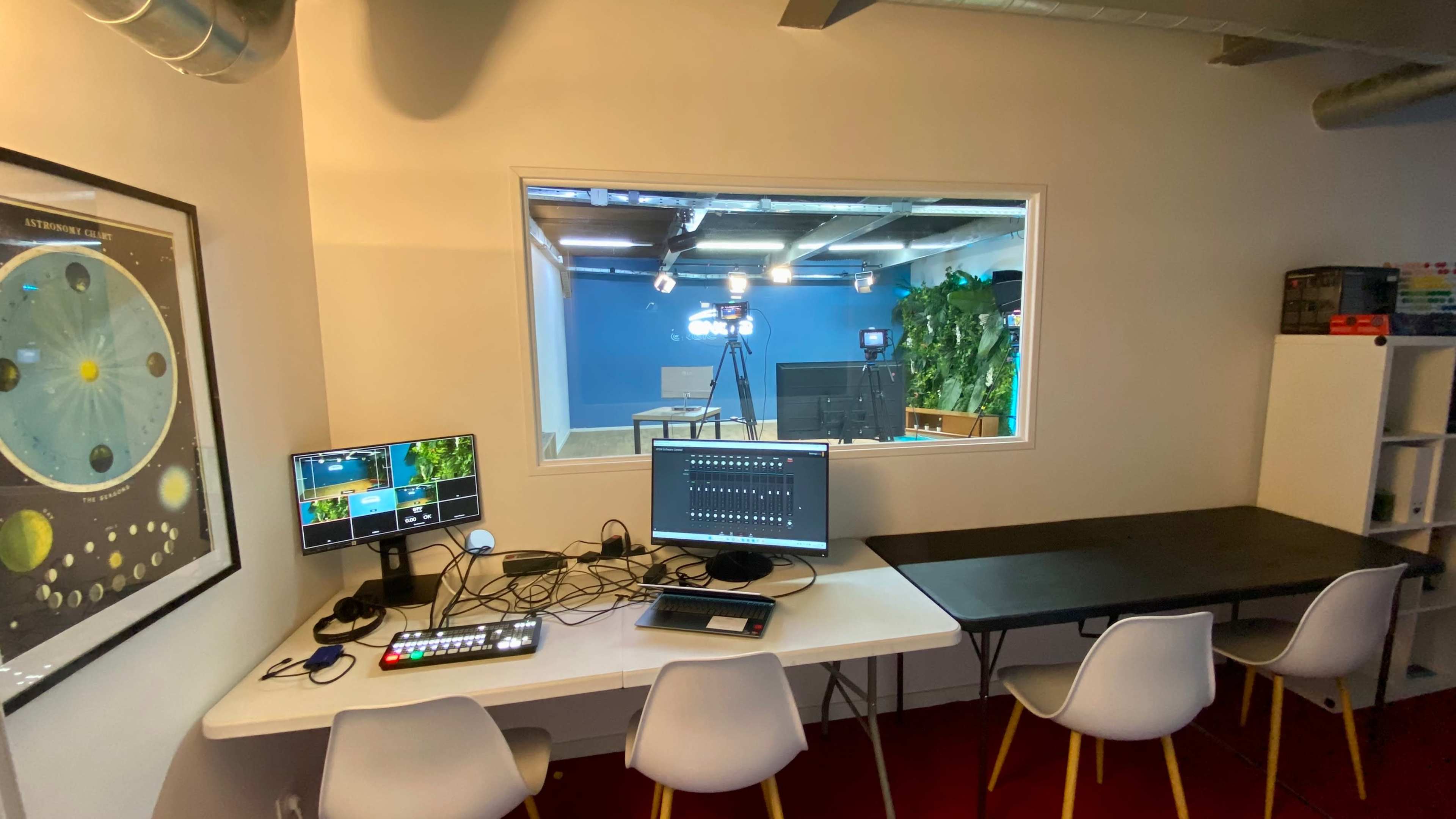 A modern workspace with a computer, video equipment, and a large window overlooking a blue studio setup with cameras and plants.