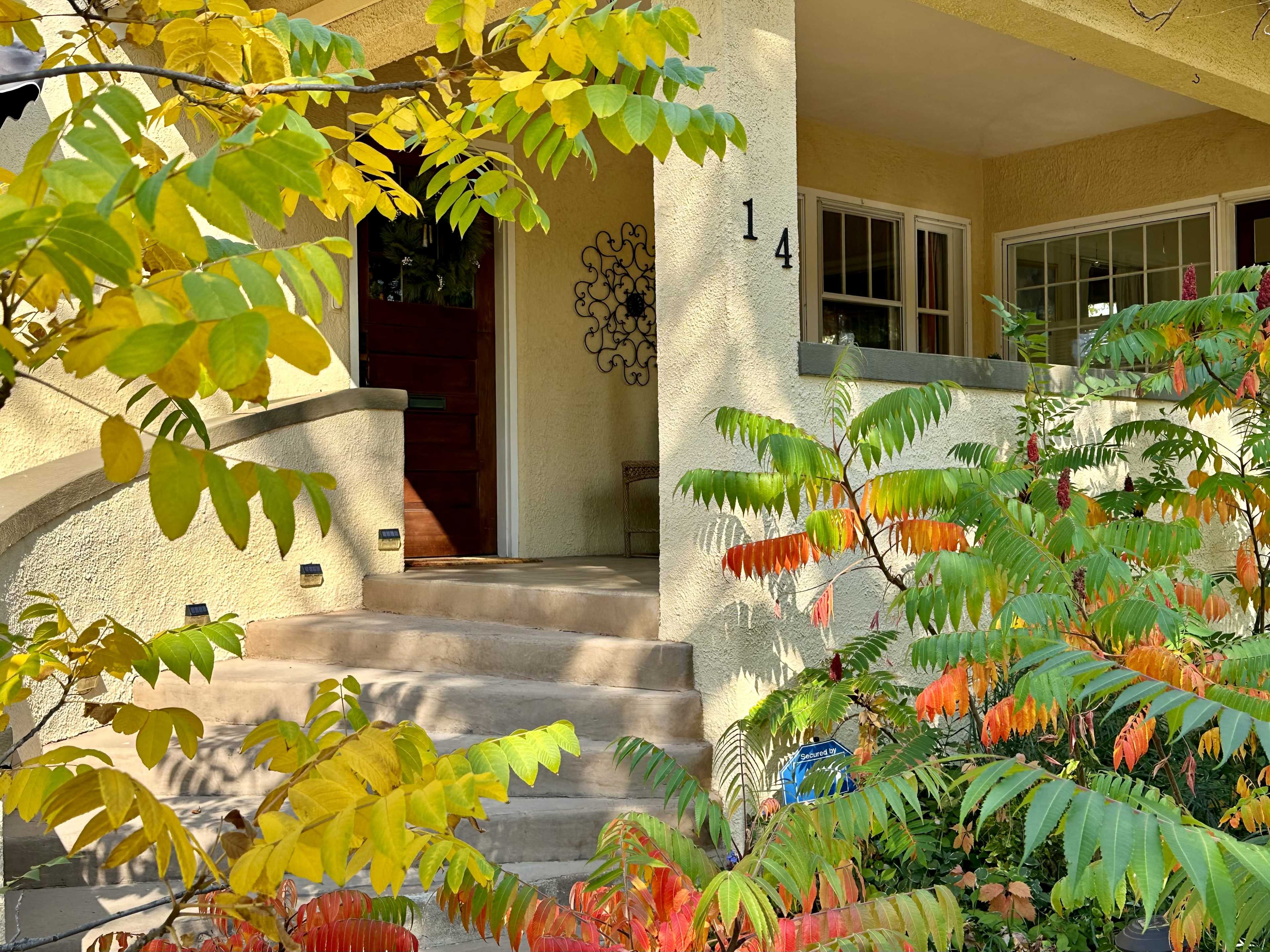 A yellow house with a prominent front porch and steps is partially obscured by colorful foliage.