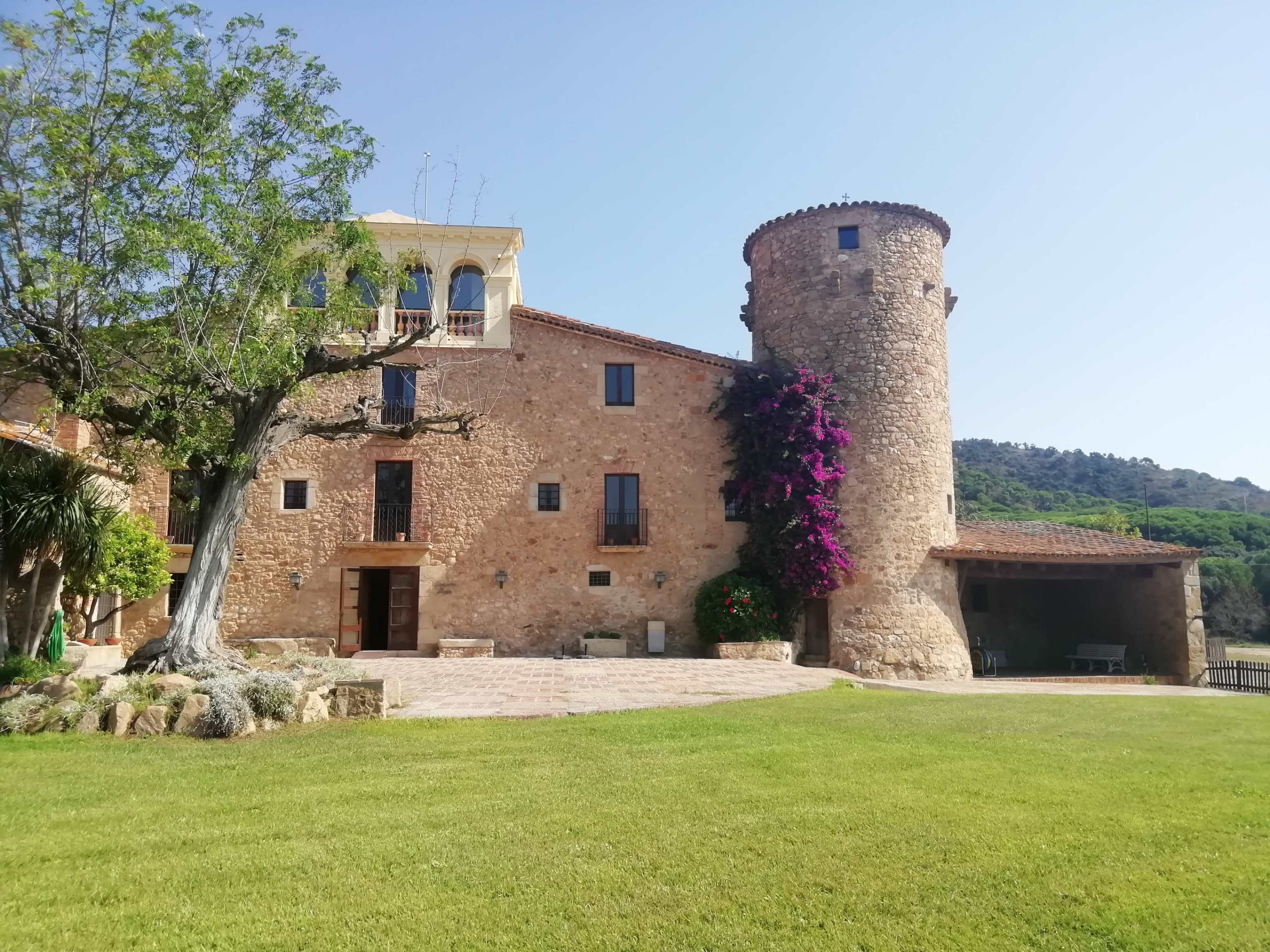 The image shows a rustic stone building with a round tower and vibrant purple flowers, set against a clear blue sky and green hills.