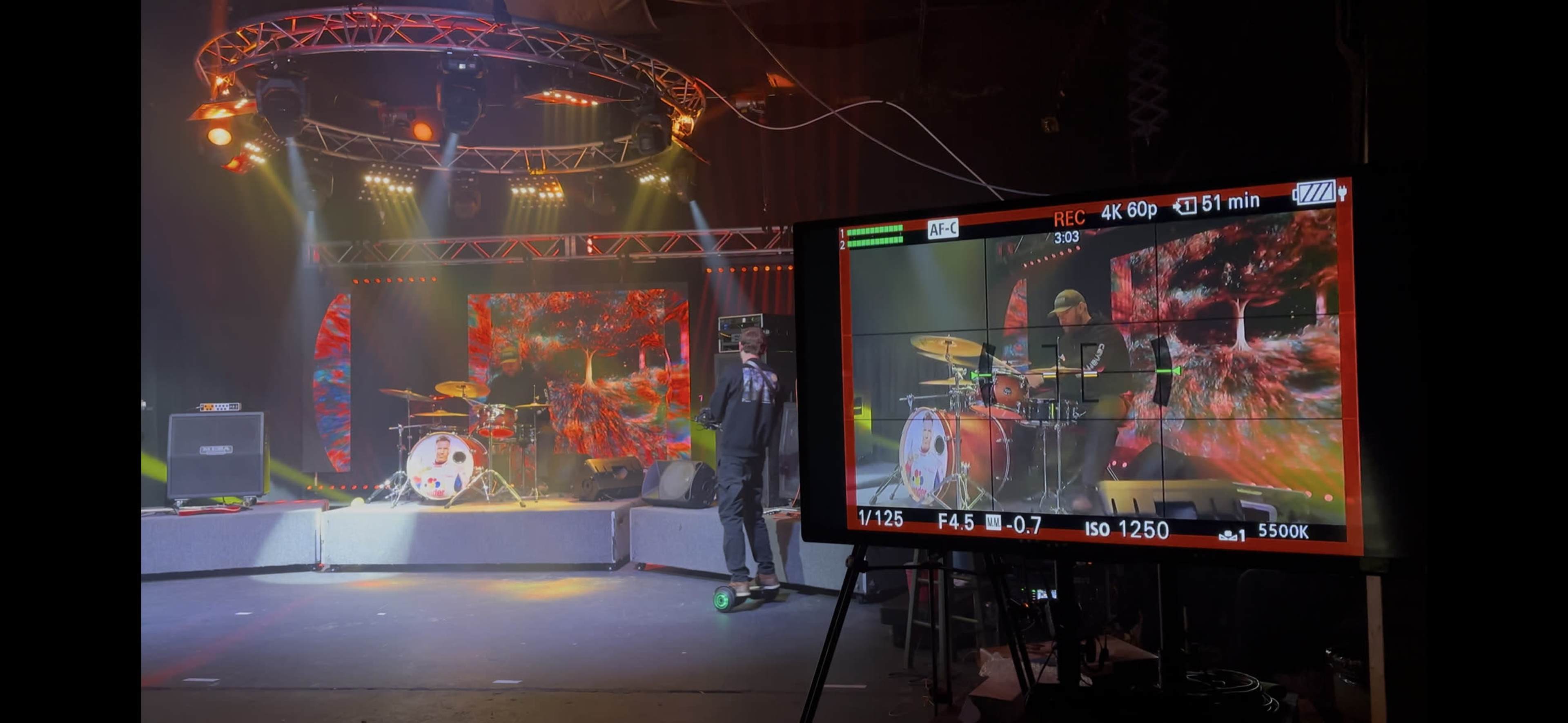 A musician performs on stage with a drum set and vibrant visual displays in the background, while a camera monitor captures the scene.