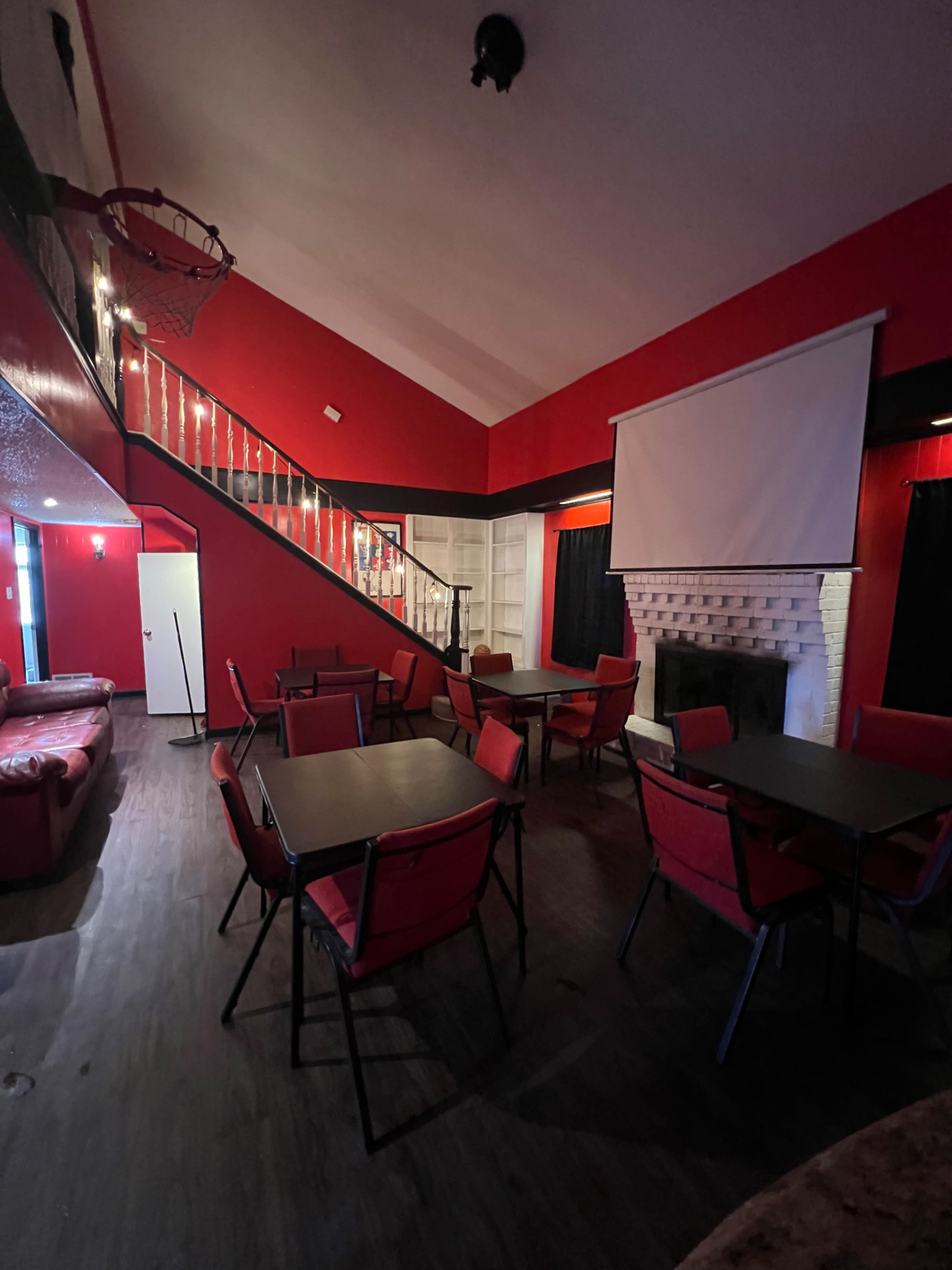 The image shows a room with red walls, a staircase, several black tables with red chairs, and a white projection screen above a fireplace.