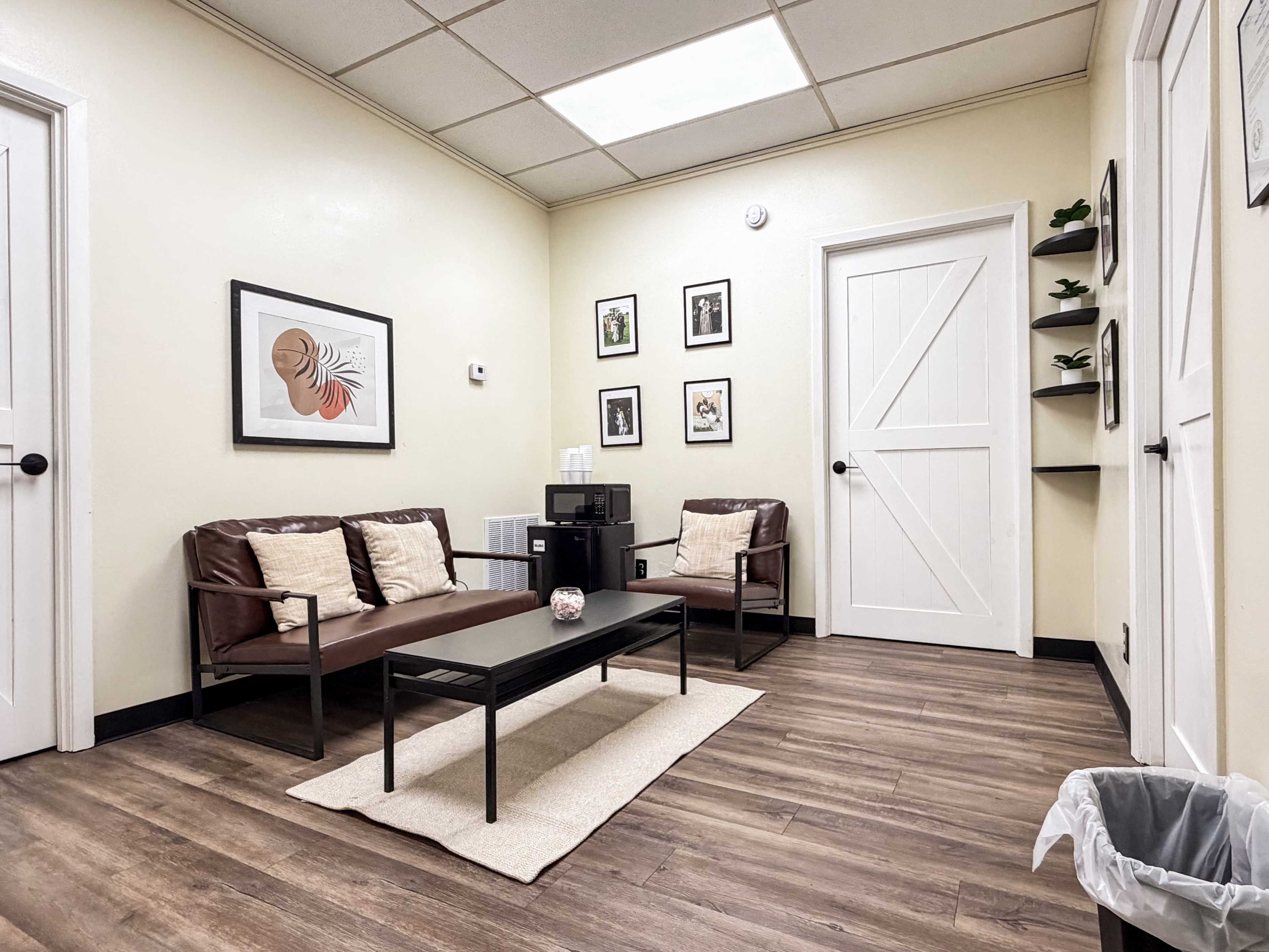 The image shows a small waiting room with two brown leather chairs, a coffee table on a rug, framed pictures on the walls, and a white door leading to another room.