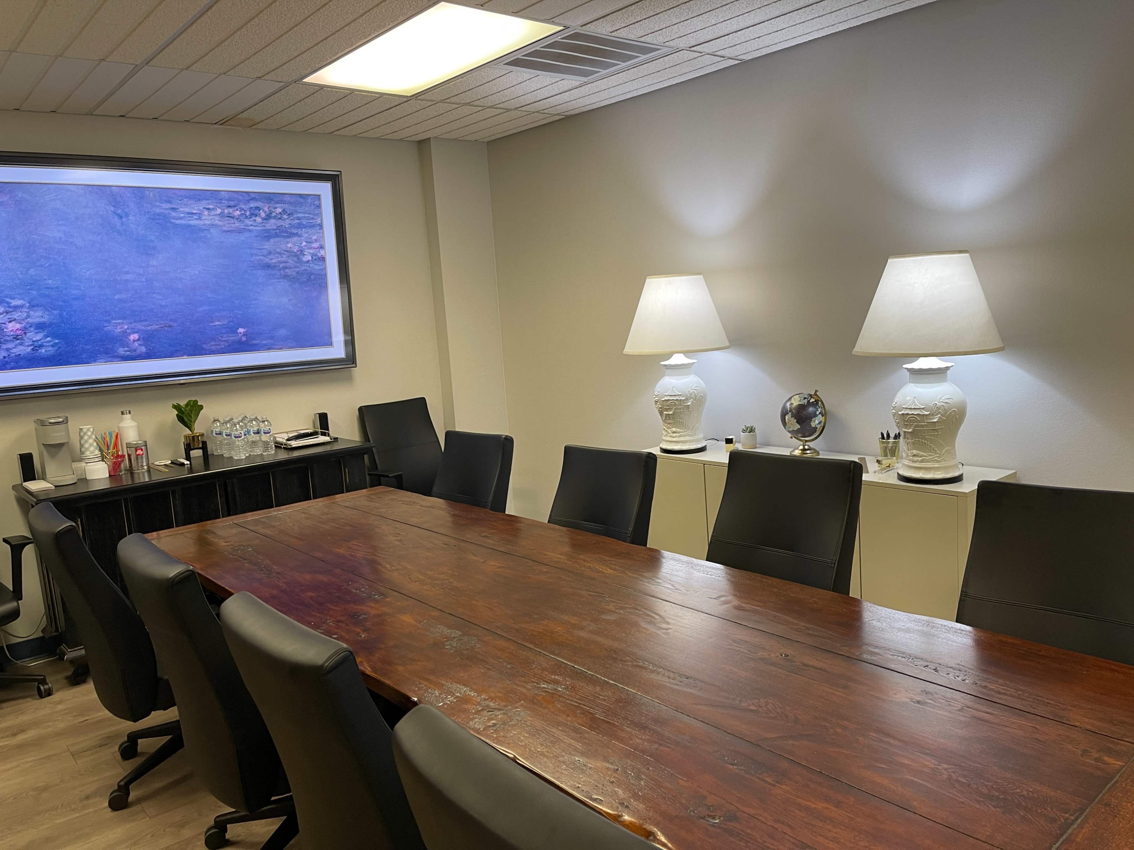A conference room with a large wooden table, black leather chairs, two white lamps, and a window displaying a painting.