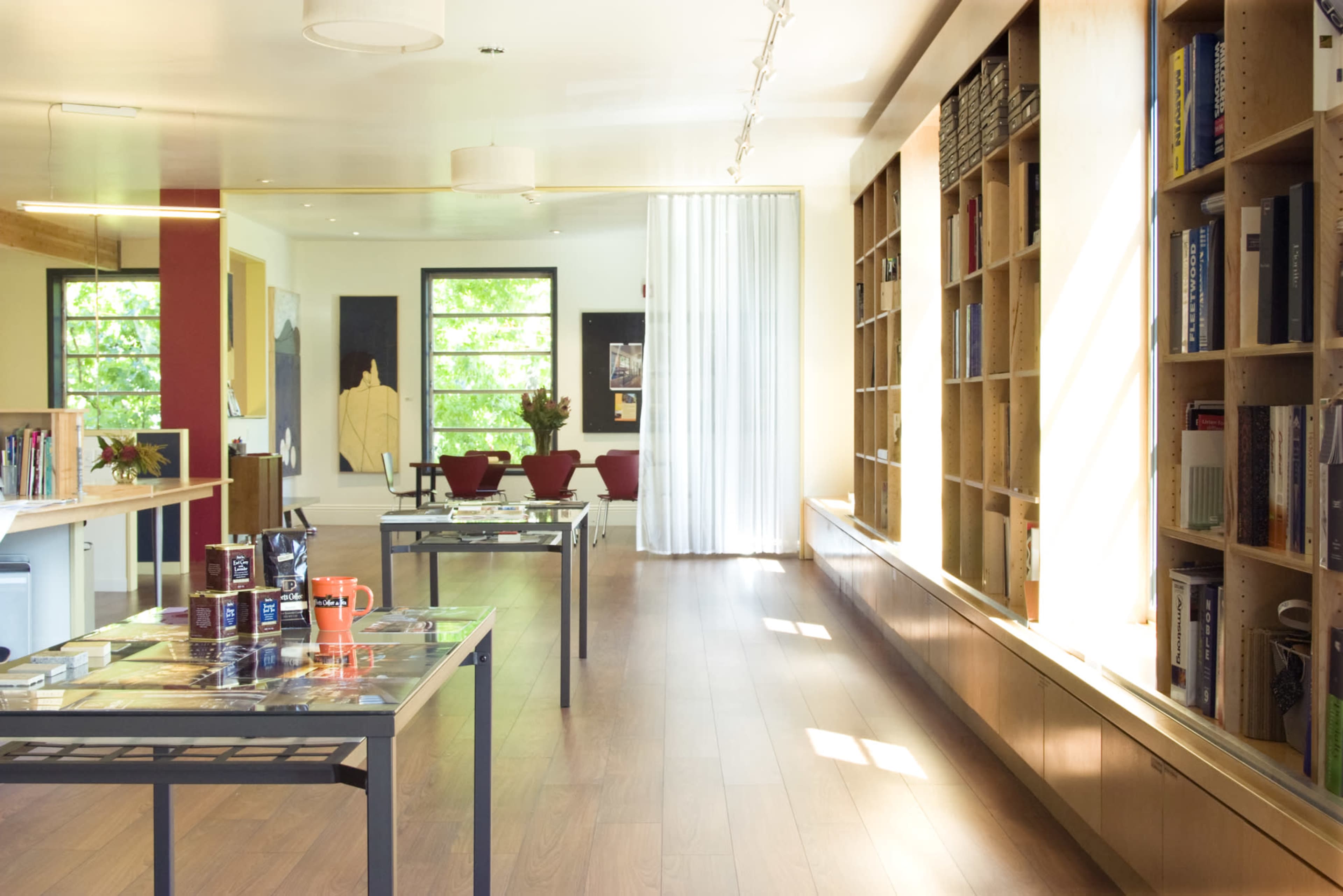 A modern interior space featuring wooden flooring, sleek furniture, shelving filled with books, and large windows allowing natural light to flood the room.