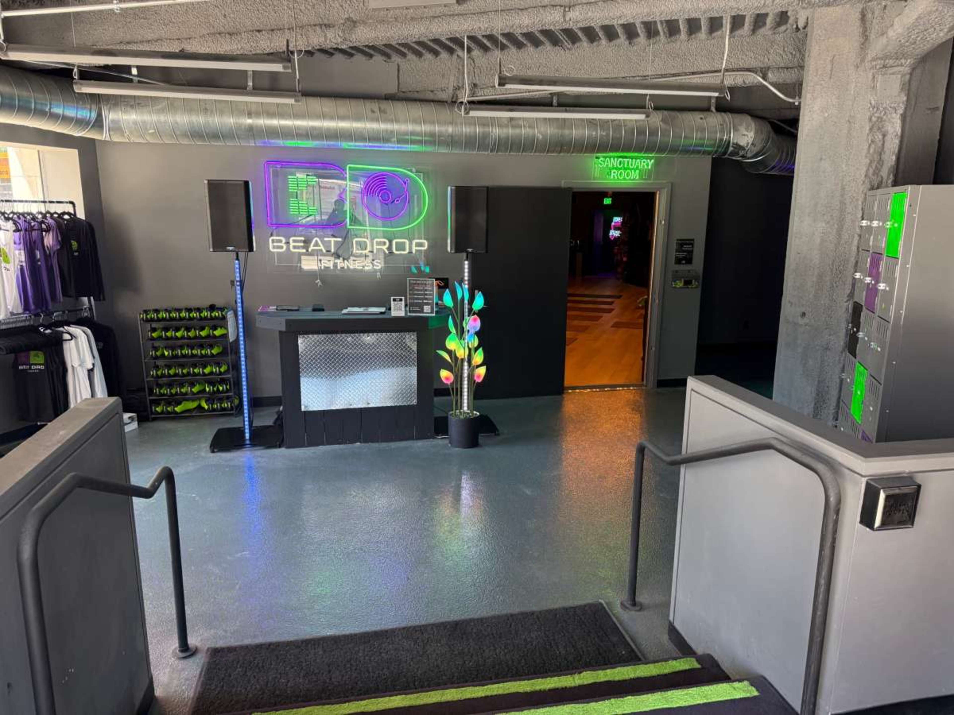 The image shows the entrance of a fitness center named "Beat Drop Fitness," featuring a reception desk, a brightly lit sign, and an adjacent room labeled "Sanctuary Room."