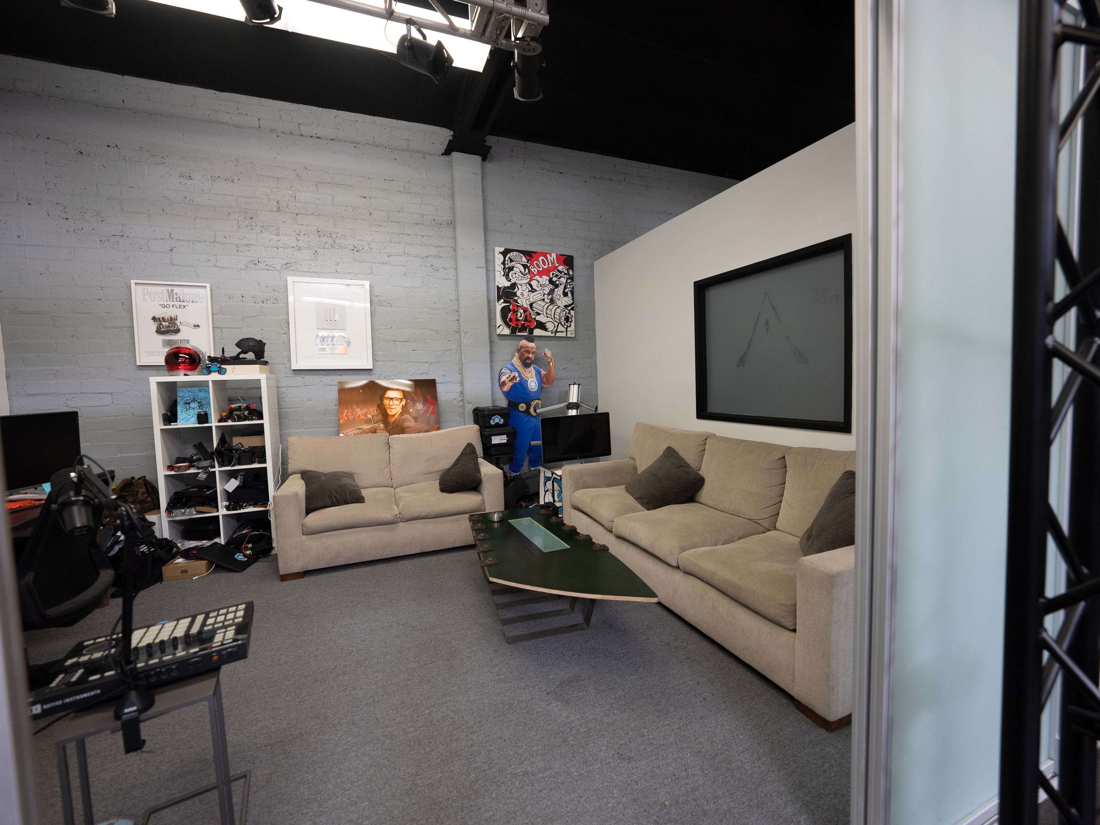 A man in a blue jumpsuit stands in a modern office space featuring two beige sofas, a coffee table, and various items on shelves.
