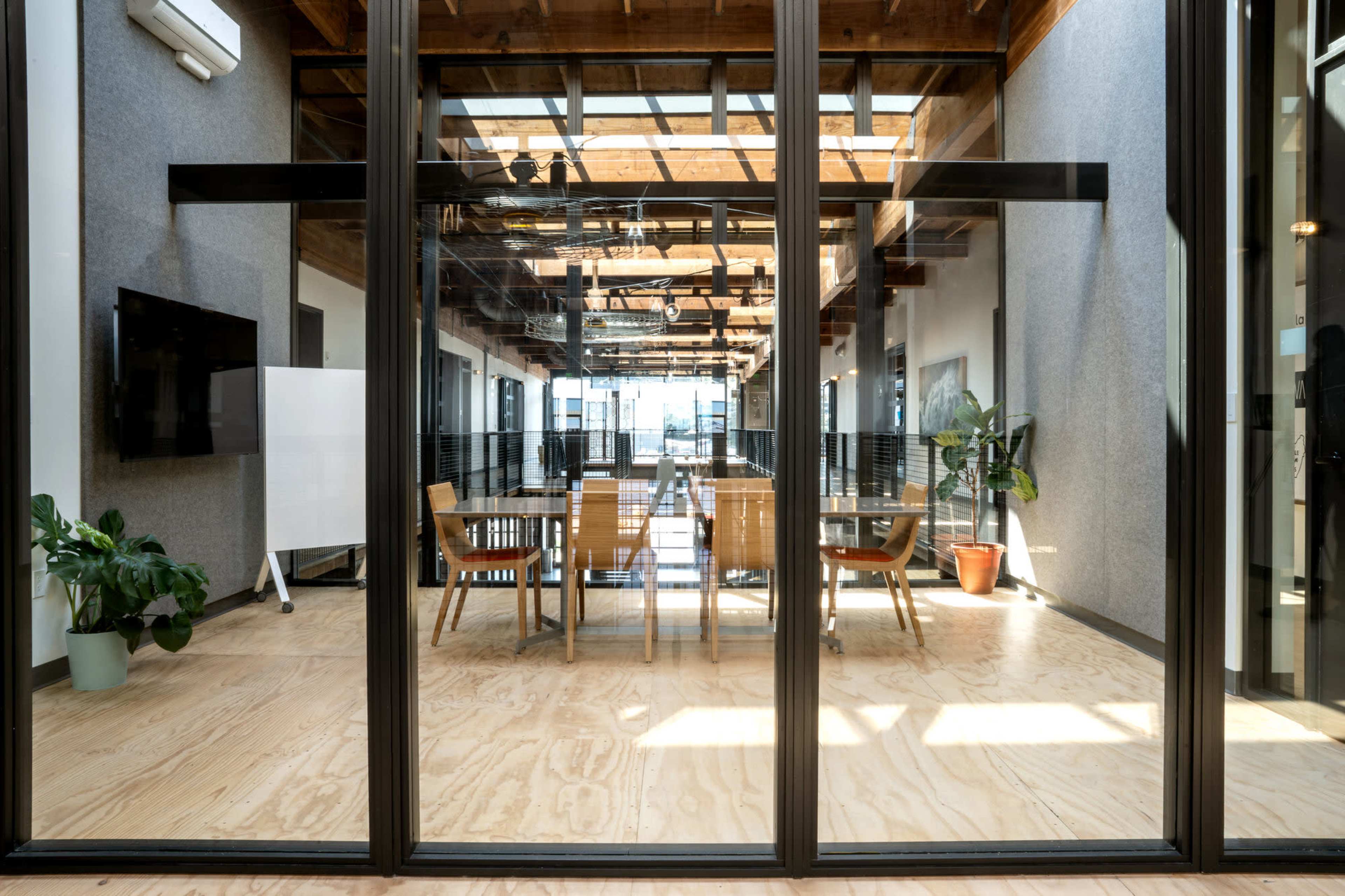 The image shows a modern conference room with a wooden table and chairs, enclosed by glass walls, and featuring plants and a large window that allows natural light to enter.