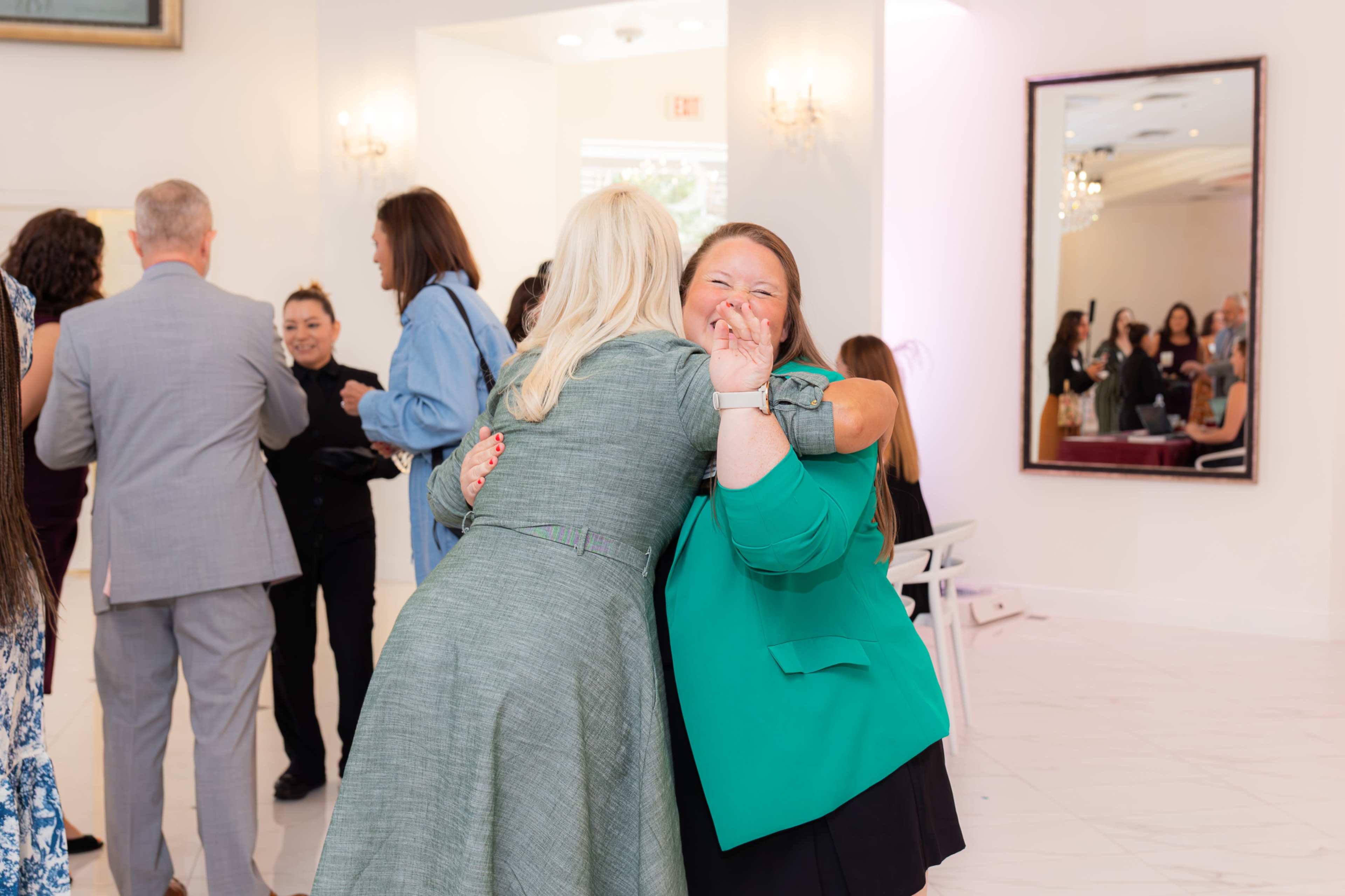 Two women embrace in a welcoming indoor event space, while other attendees chat and mingle in the background.
