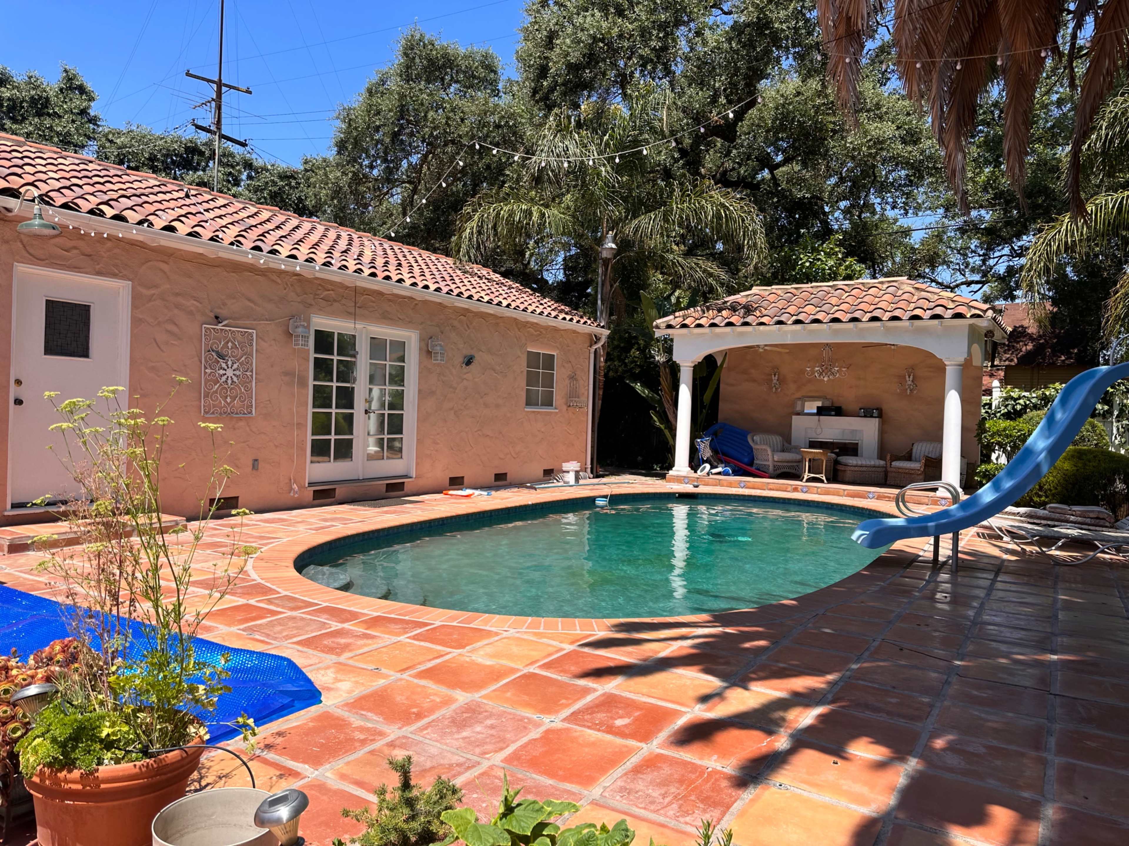 The image shows a backyard pool surrounded by a tiled patio, with a slide, a small gazebo, and a house with a textured wall.