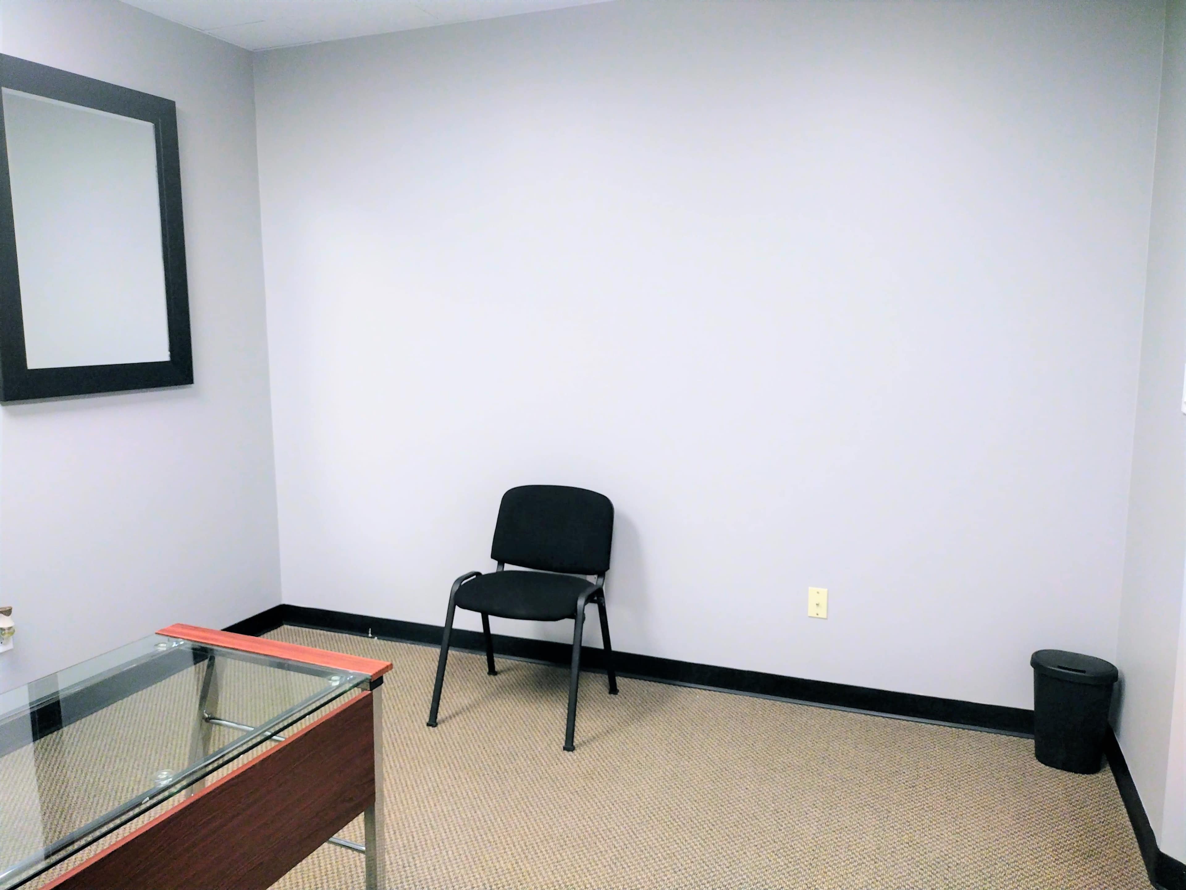 A small, empty room with a gray wall, a black chair, a glass-top desk, and a black trash can.