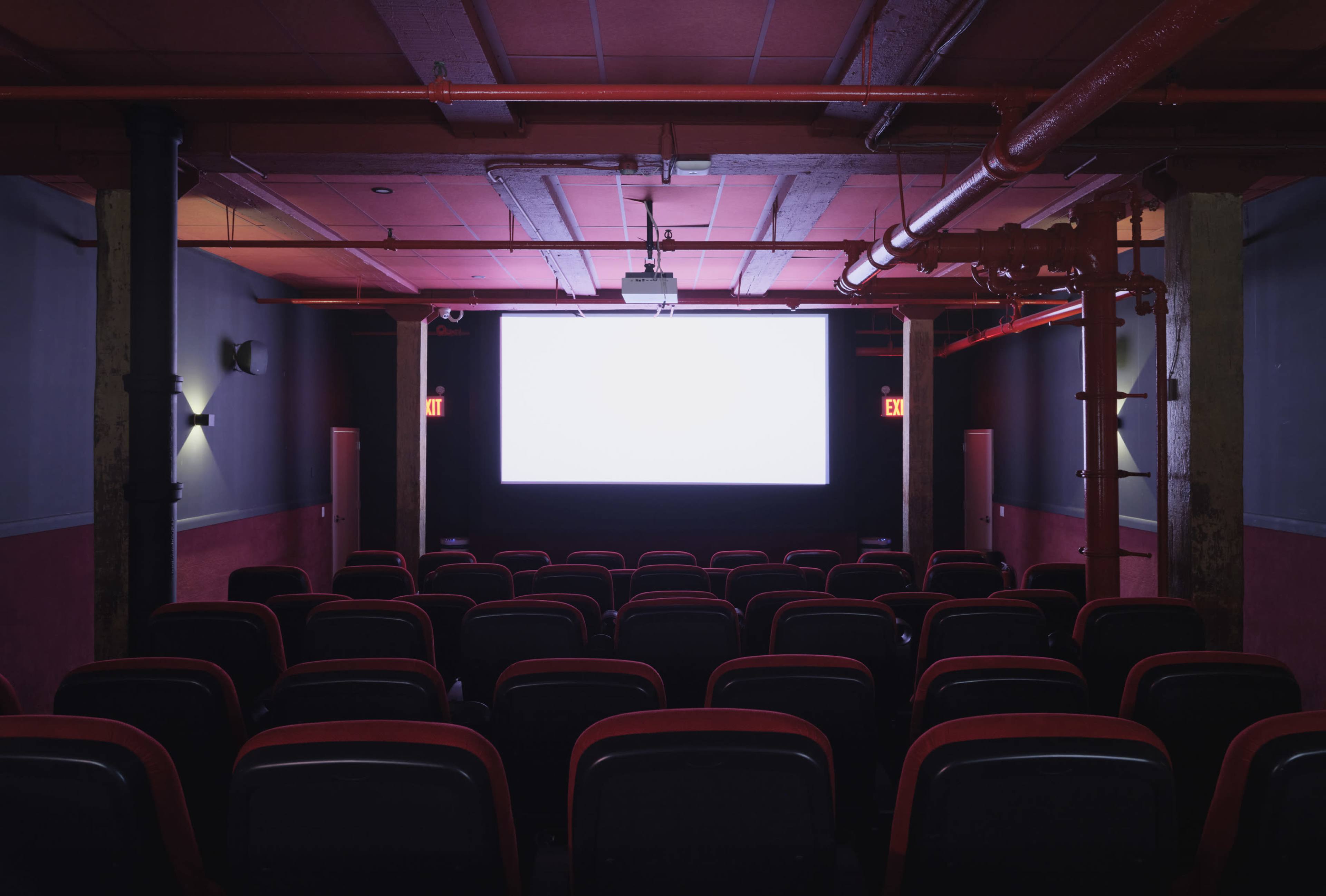 The image shows an empty movie theater with red chairs facing a large blank screen.