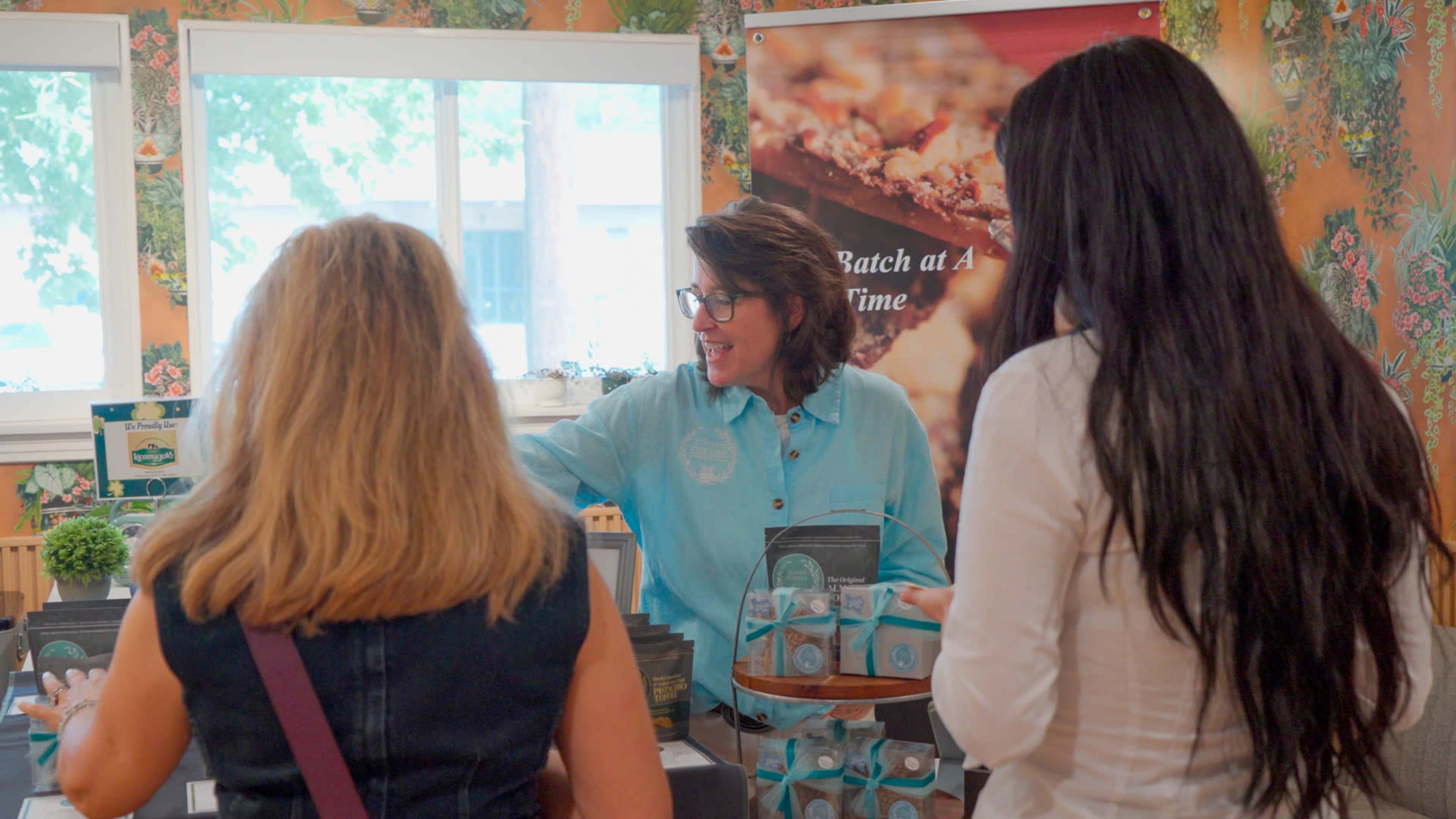 A woman in a blue shirt speaks with two customers at a table displaying various products in a shop adorned with floral wallpaper.