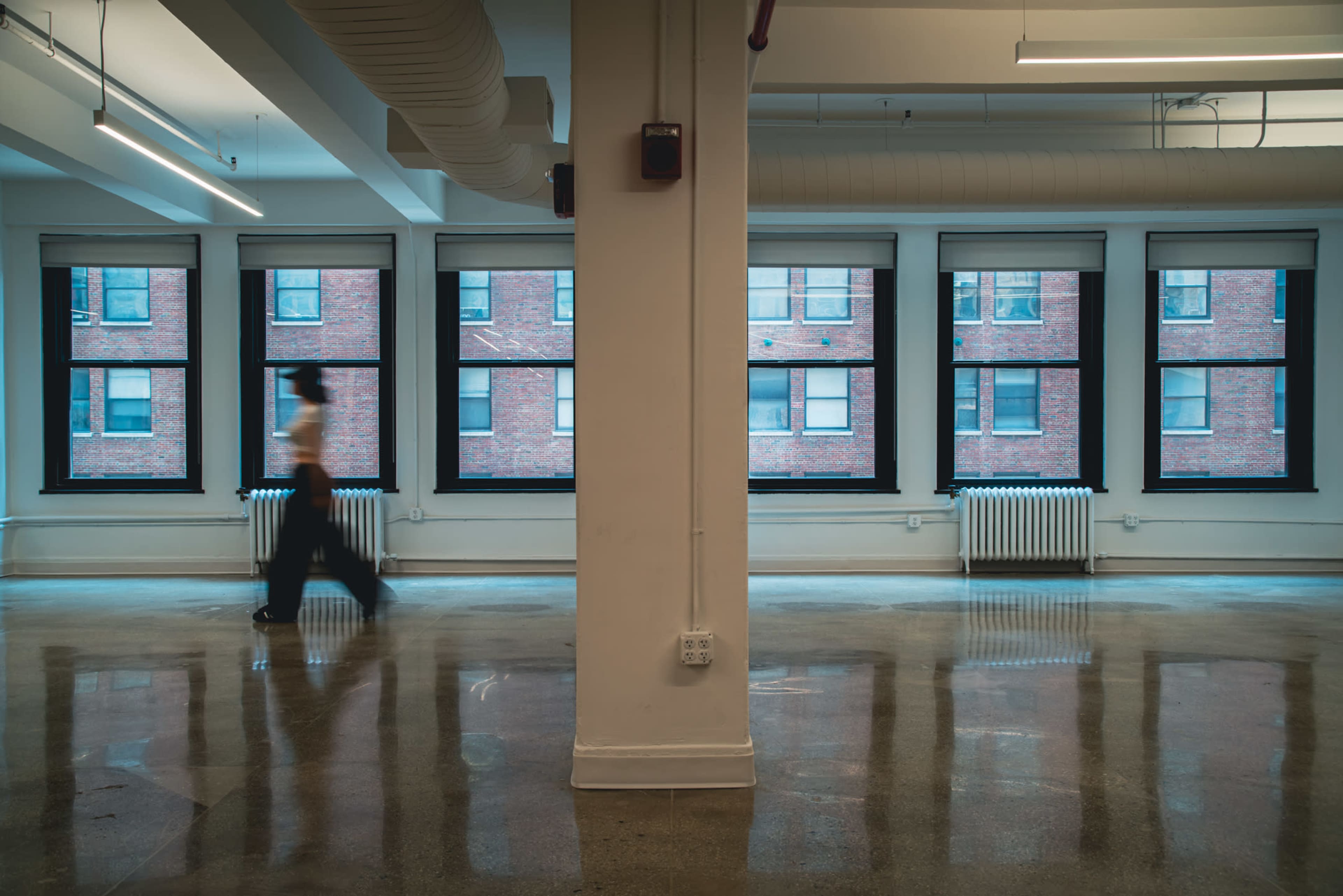 A person walks through a spacious, empty room with large windows and polished concrete floors.