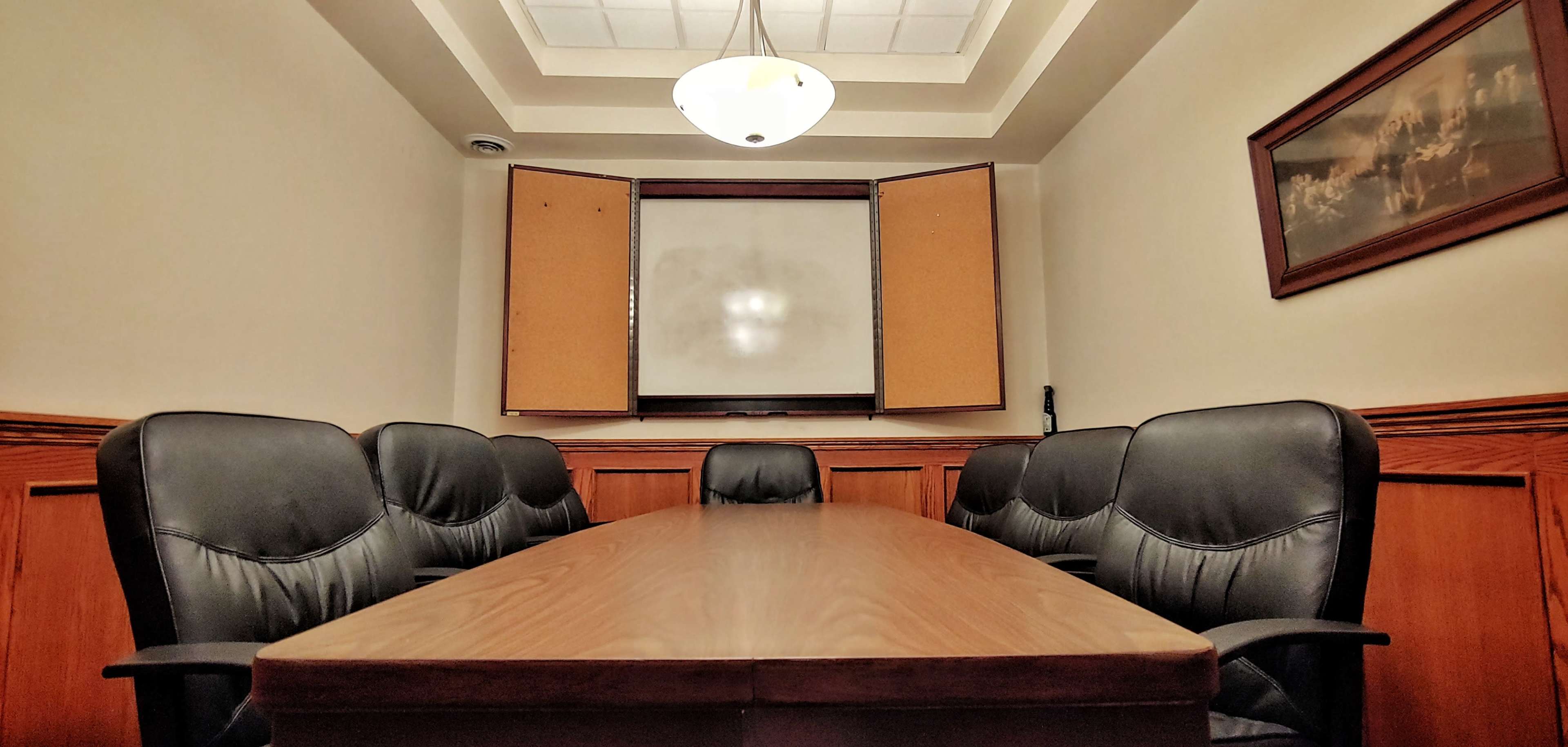 A conference room features a long wooden table surrounded by black leather chairs, with a whiteboard at one end and windows partially open.