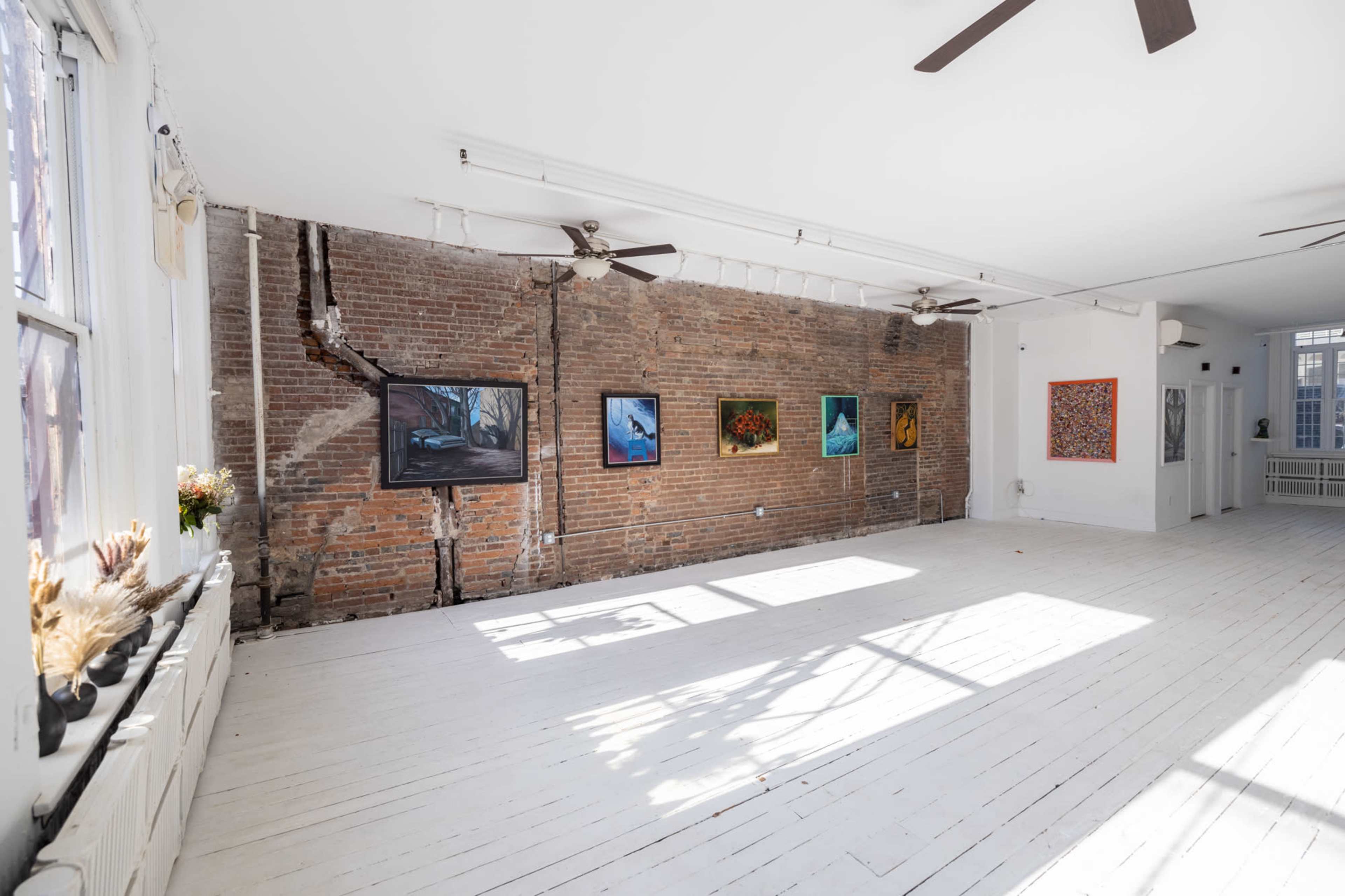An art gallery with exposed brick walls and white wooden floors, displaying various framed paintings.