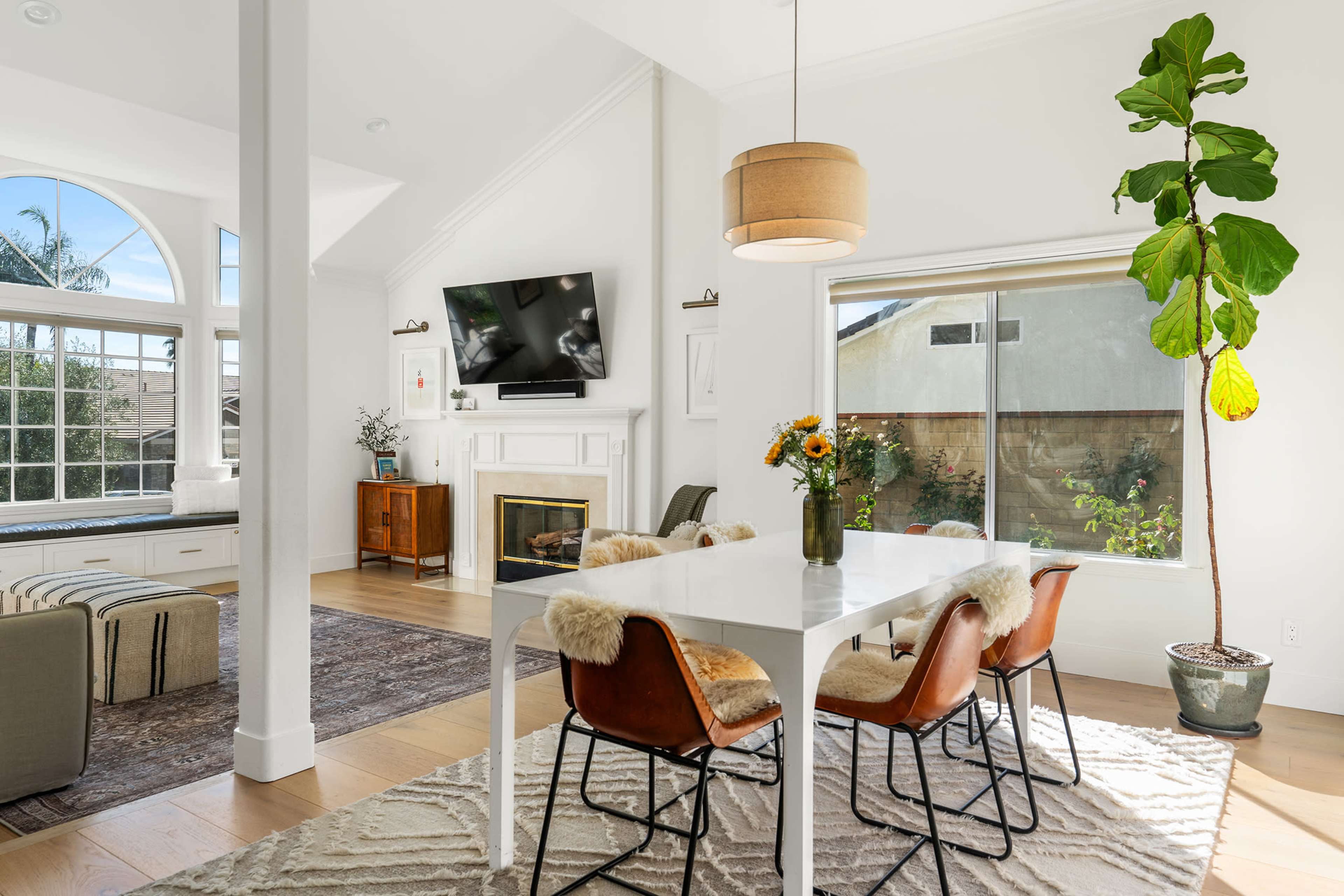 A bright dining area features a white table surrounded by chairs, with a vase of flowers and a large window overlooking greenery.