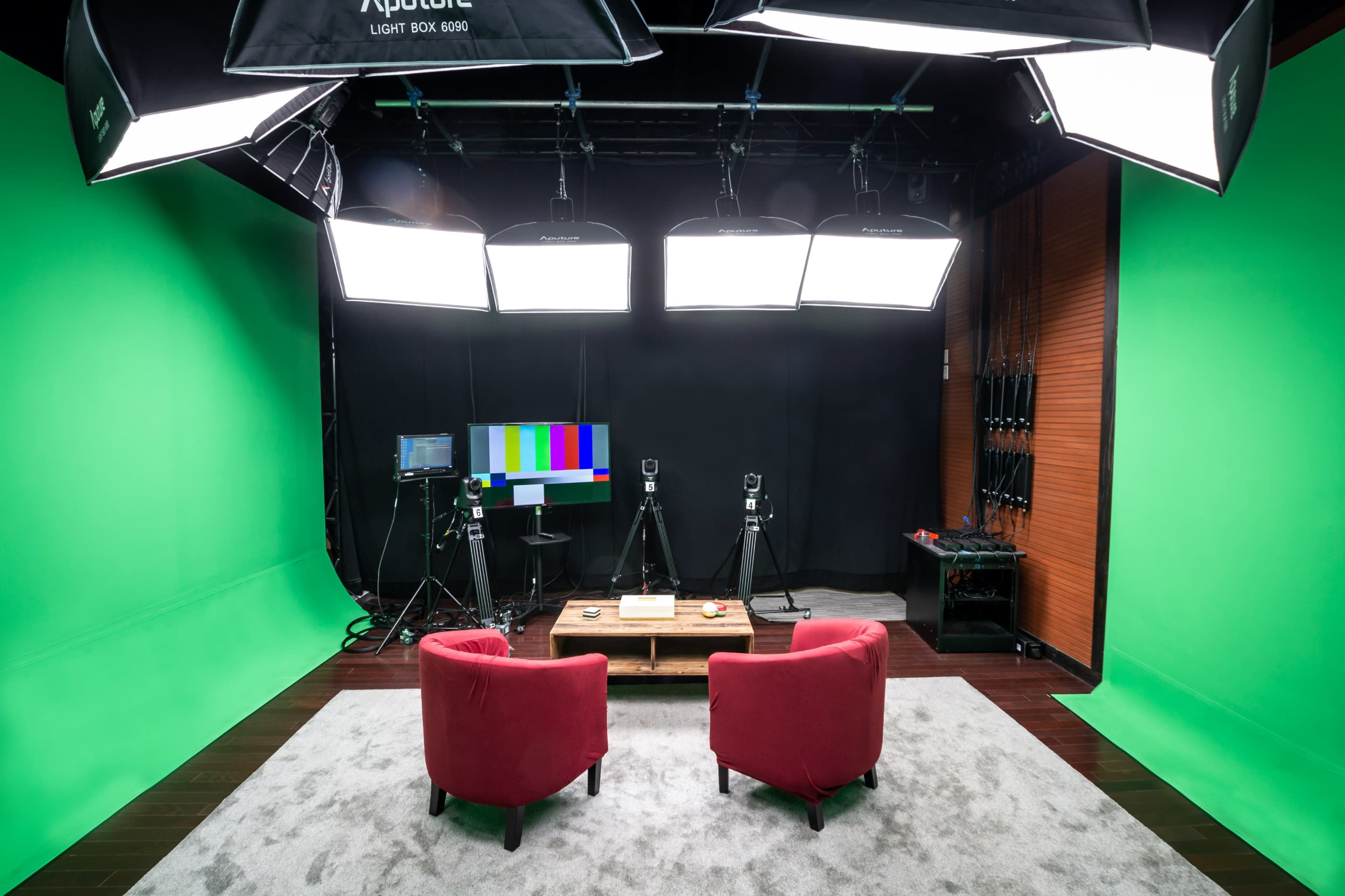 A video recording studio with two red chairs facing a wooden table, surrounded by green and black backdrop panels and equipped with multiple lights and cameras.
