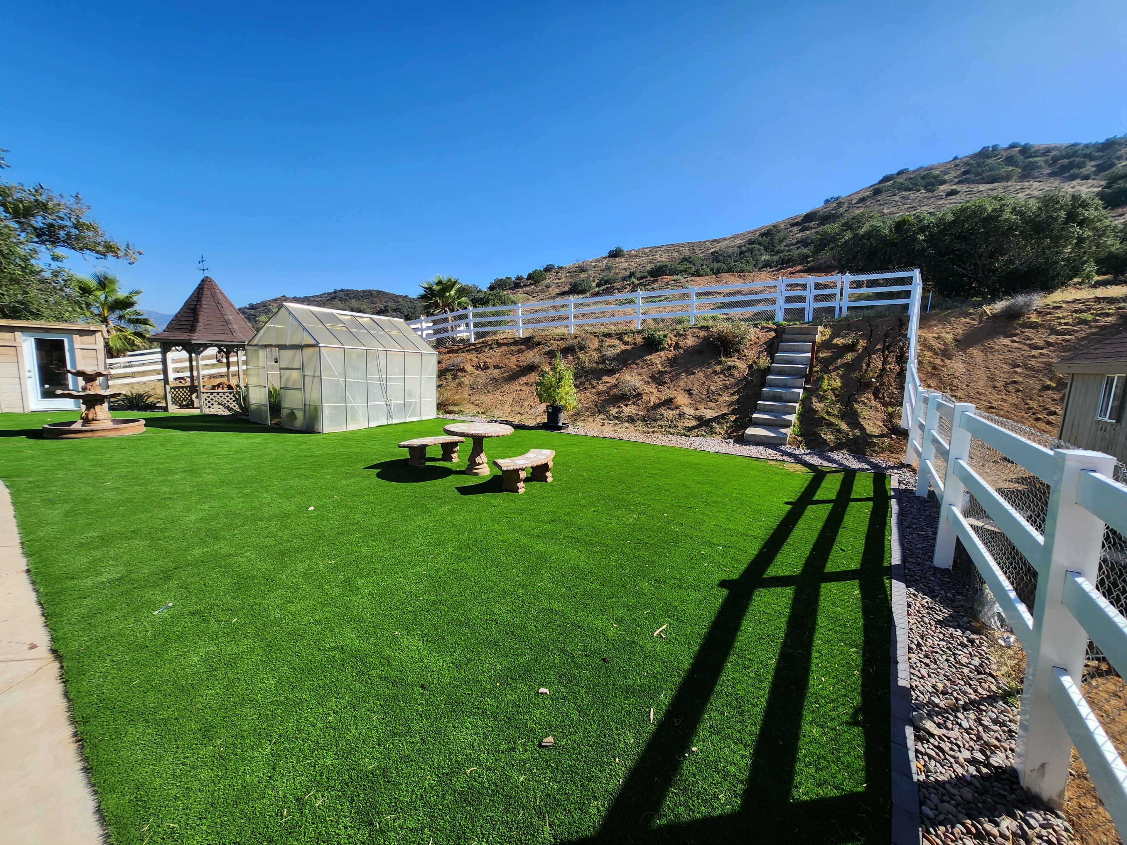 A landscaped area featuring artificial grass, a greenhouse, and a white fence, surrounded by a hillside.
