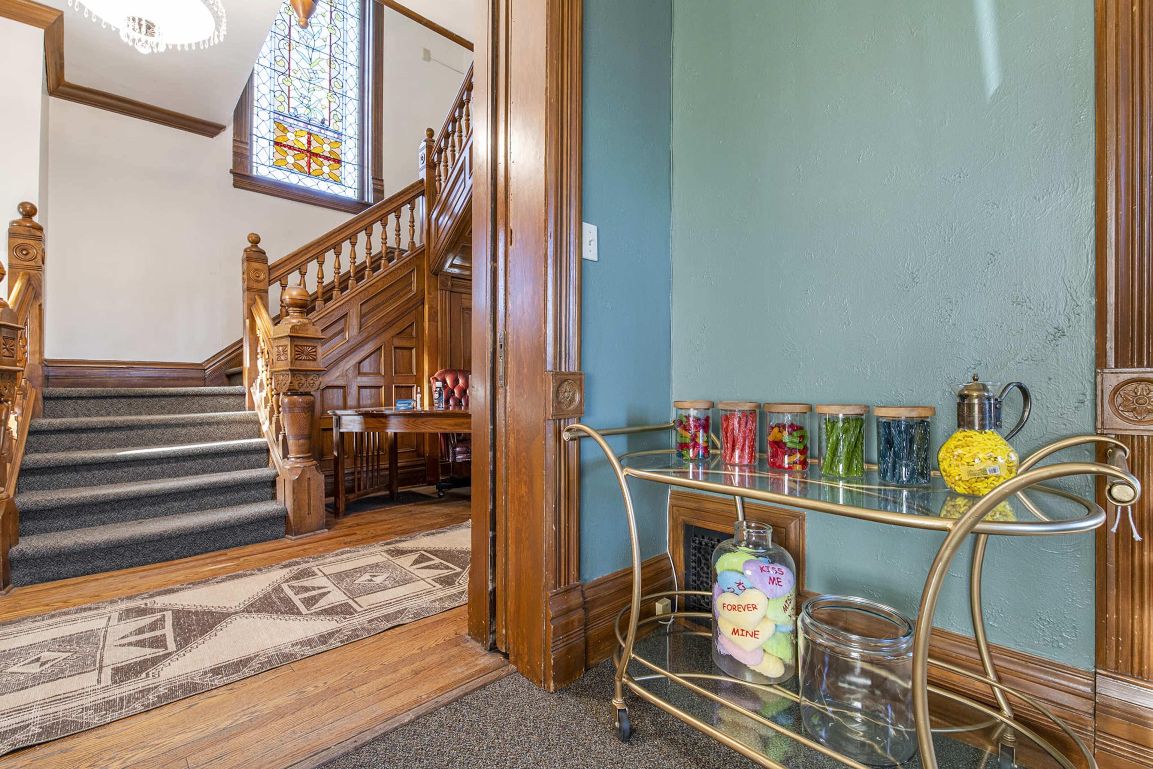 The image shows a hallway with a staircase, a wooden banister, and a decorated cart featuring jars of colored items.