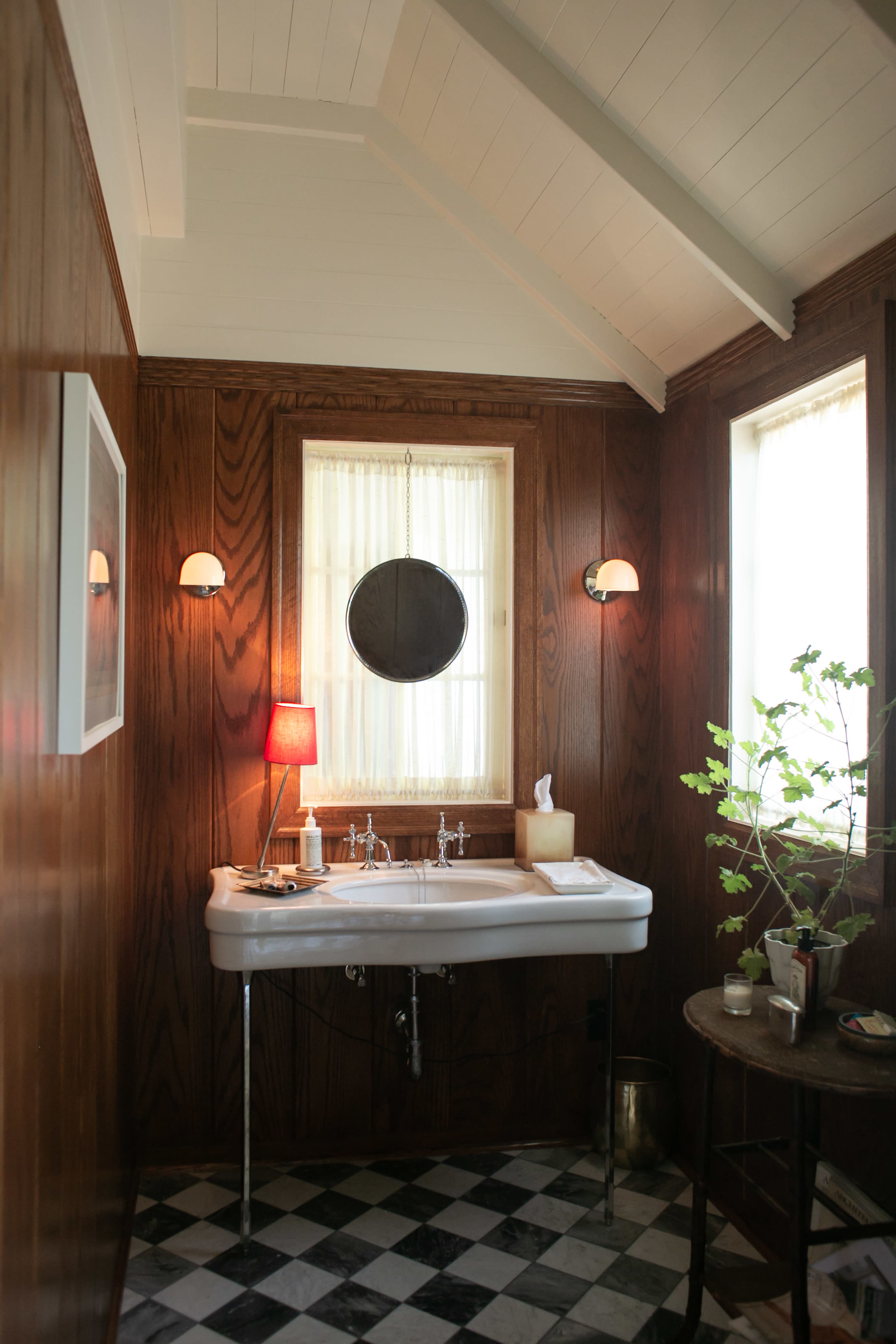 The image shows a small bathroom with a wooden paneled interior, featuring a double sink, a round mirror, and a small plant on a side table.
