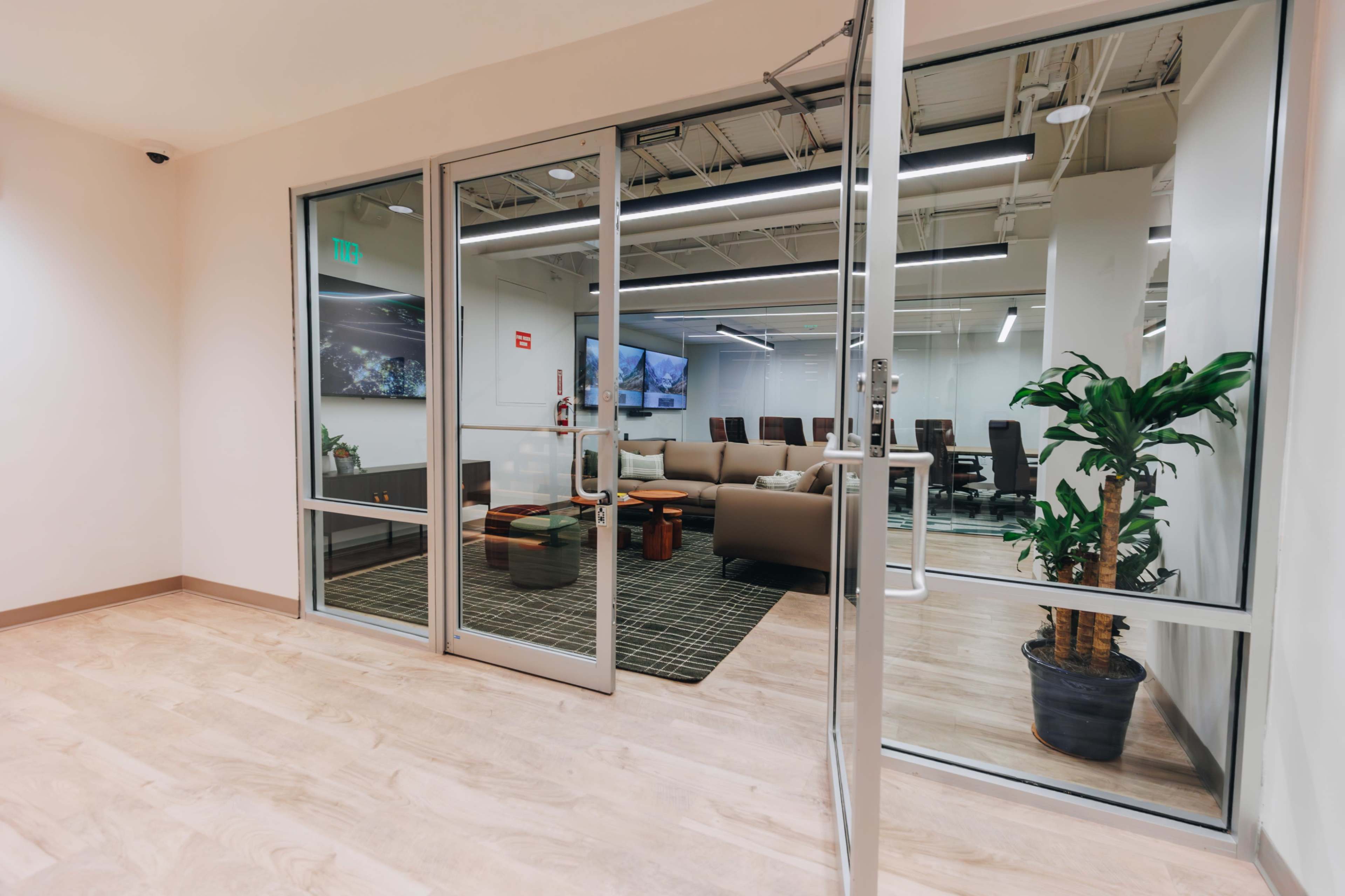The image shows a glass-walled conference room with a seating area, visible through double doors, featuring a couch, armchairs, and a large screen on the wall.