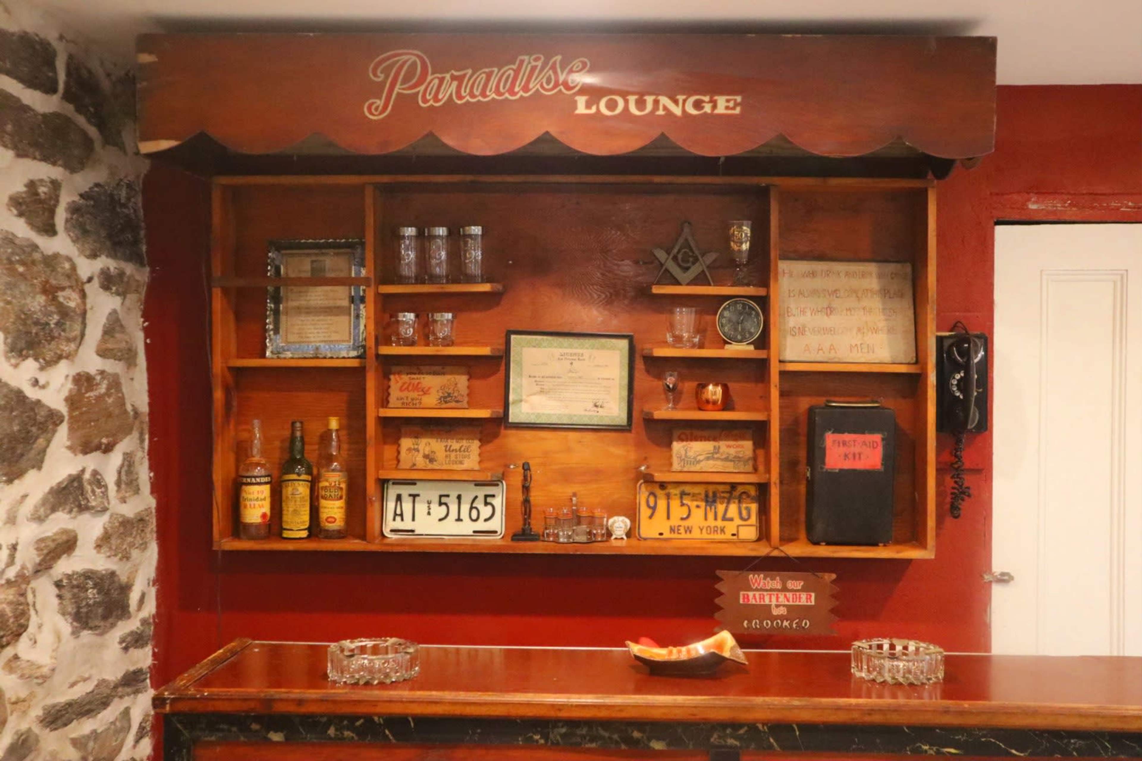 A wooden bar area with shelves displaying various bottles, several framed items, and a vintage phone, all against a stone wall backdrop.