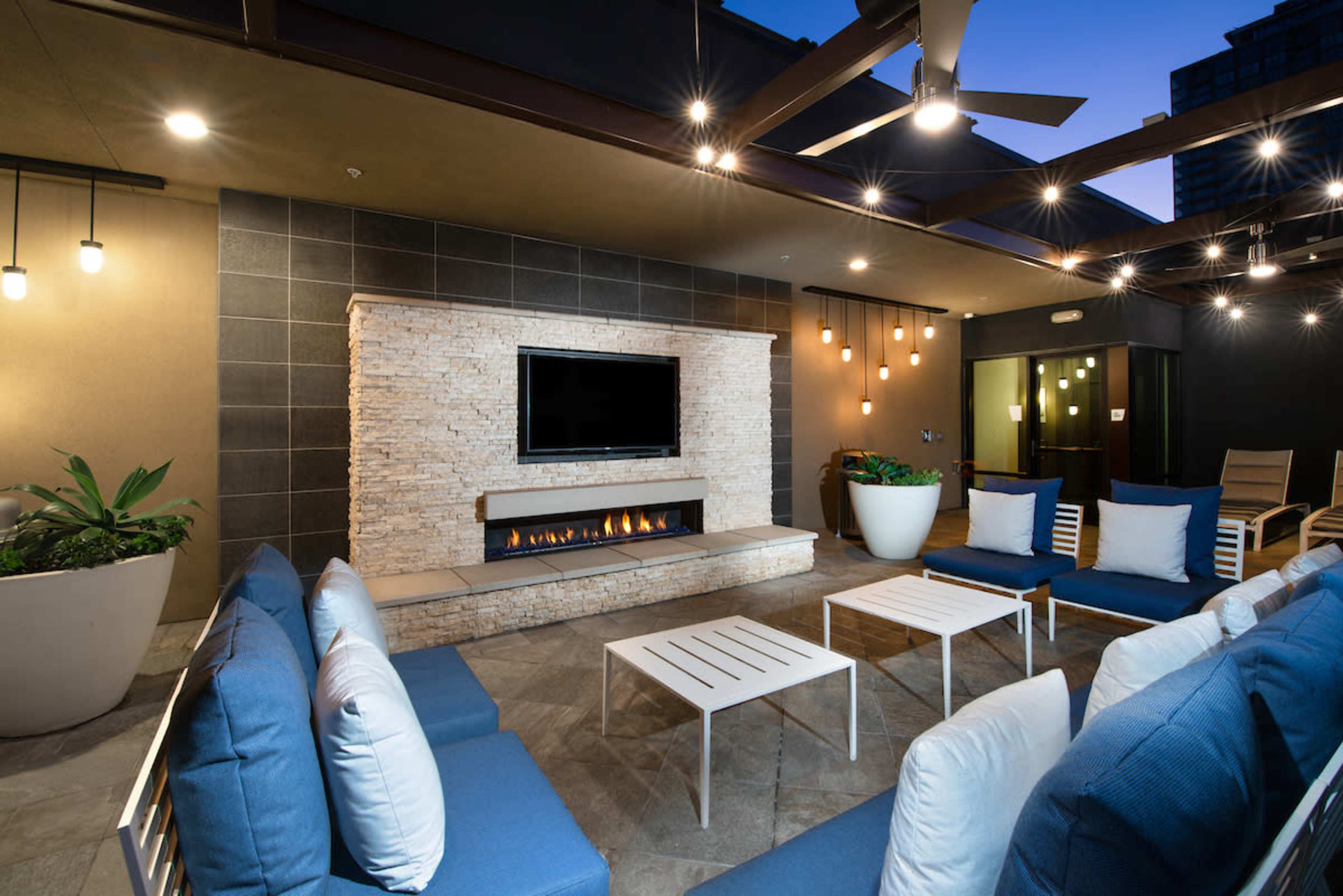 A modern outdoor lounge area features a stone fireplace, a flat-screen TV, blue seating, and decorative lighting.