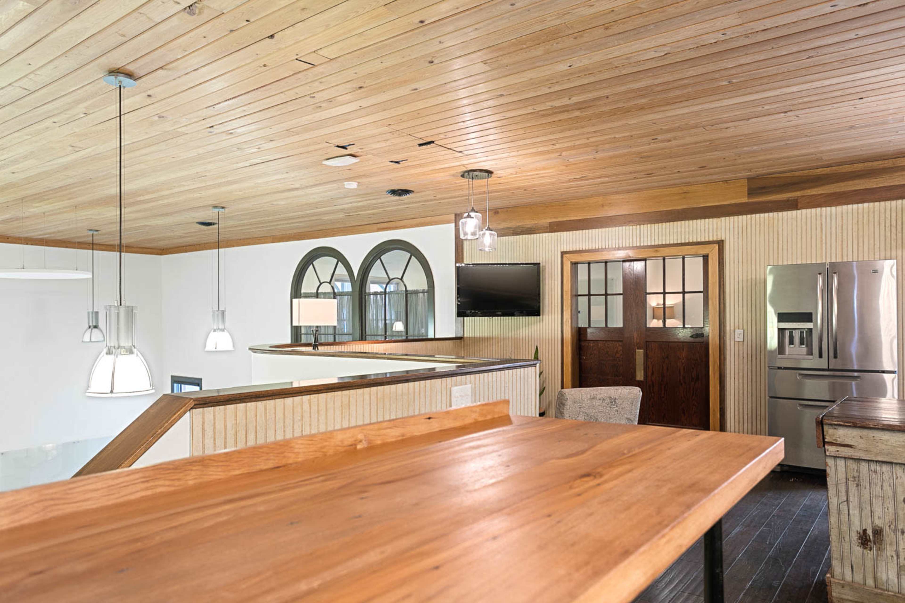 The image shows a modern interior space featuring wooden ceilings, a wooden countertop, a set of pendant lights, an arched window, and a kitchen area with stainless steel appliances.