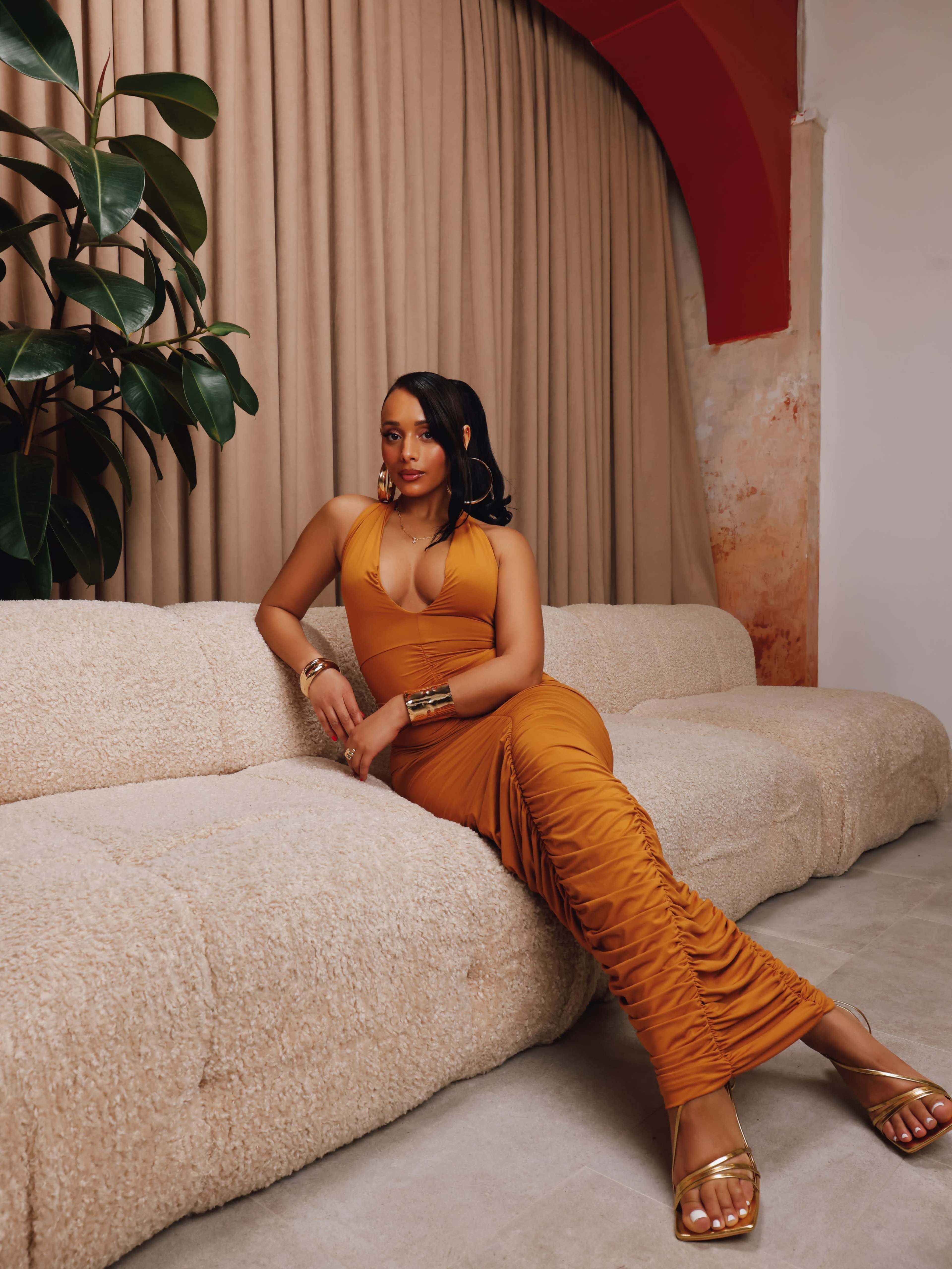 A woman in a form-fitting, rust-colored dress sits on a beige, textured couch against a backdrop of draped curtains and a potted plant.