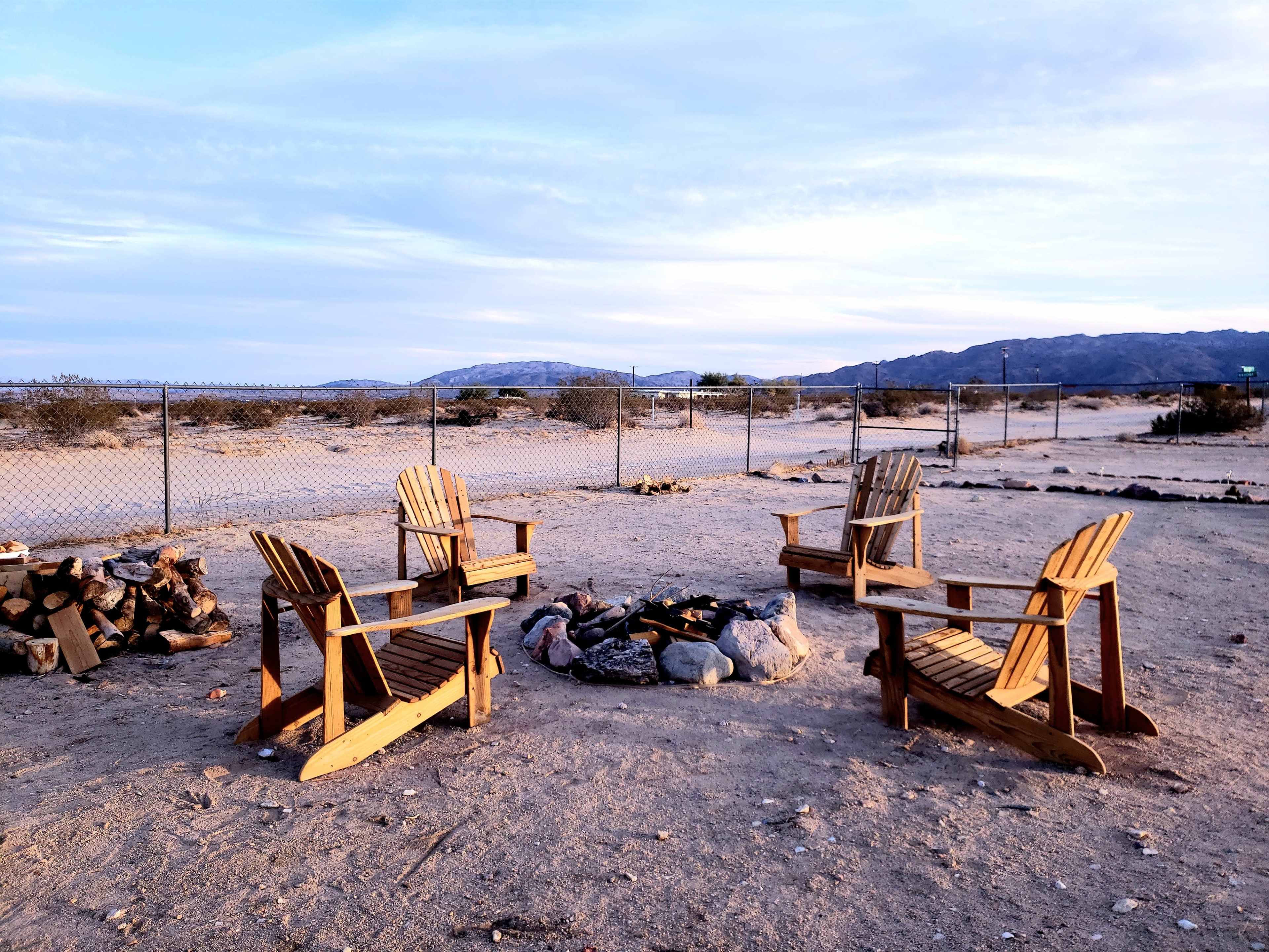 Four wooden chairs are arranged in a circle around a fire pit in a sandy outdoor area surrounded by a fence and desert landscape.