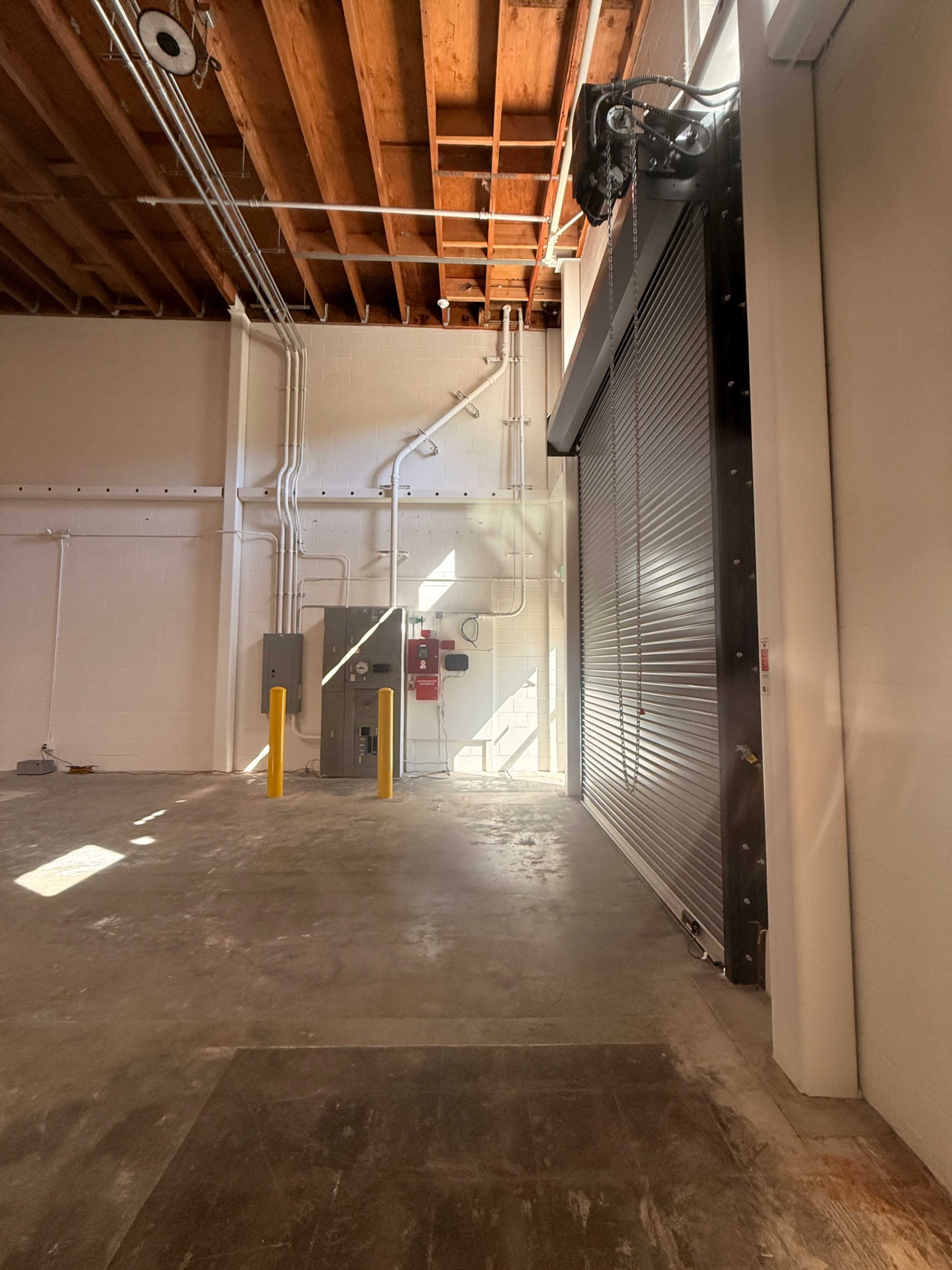 The image shows an empty industrial space featuring a roll-up door, exposed wooden beams, and utility equipment along the walls.