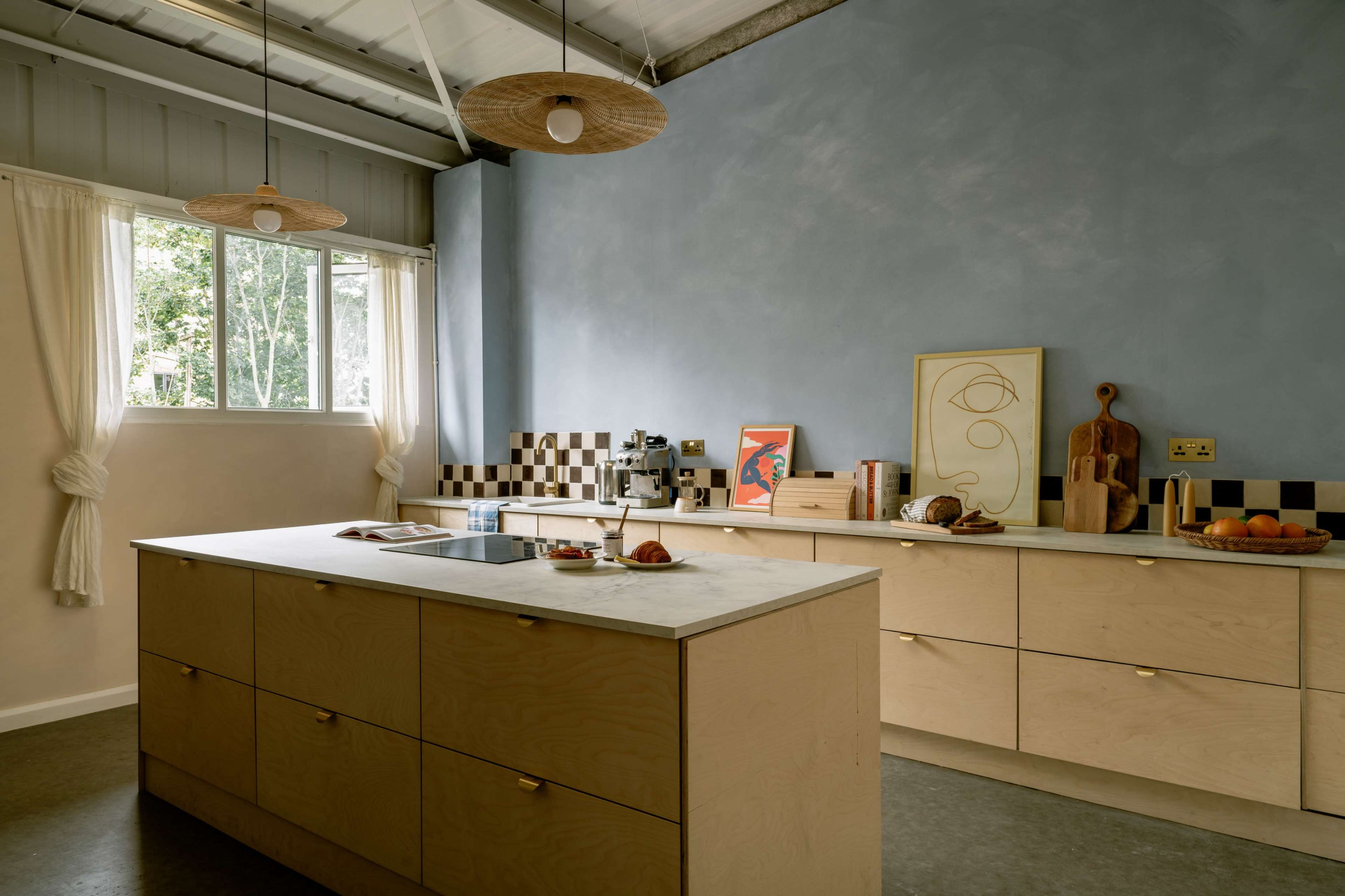 A modern kitchen with a light wooden island and cabinetry, a marble countertop, and a wall adorned with checkered tiles and artwork.
